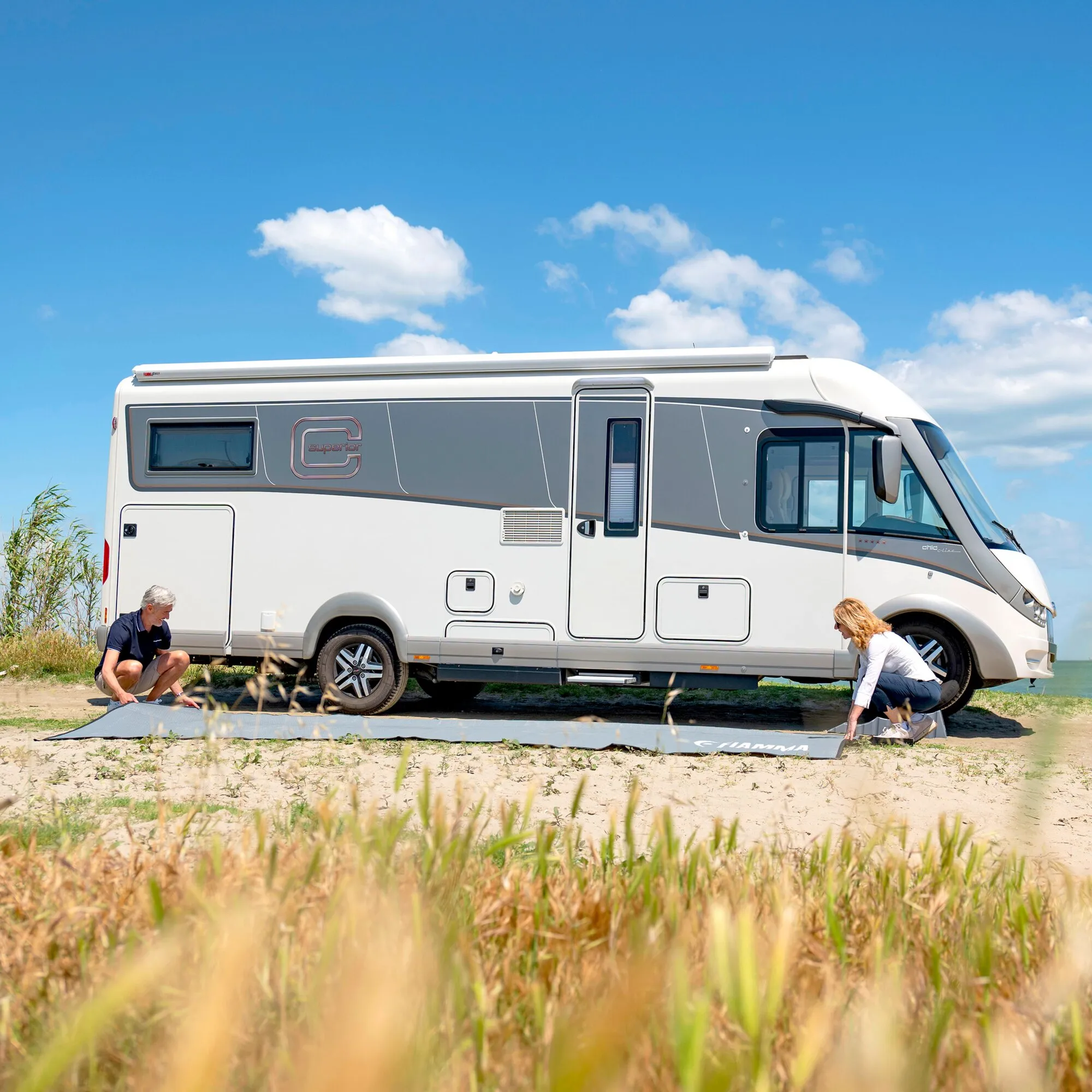 Grauer Zeltteppich Patio-Mat mit Fiamma-Logo wird vor einem weißen Wohnmobil auf Sand ausgerollt