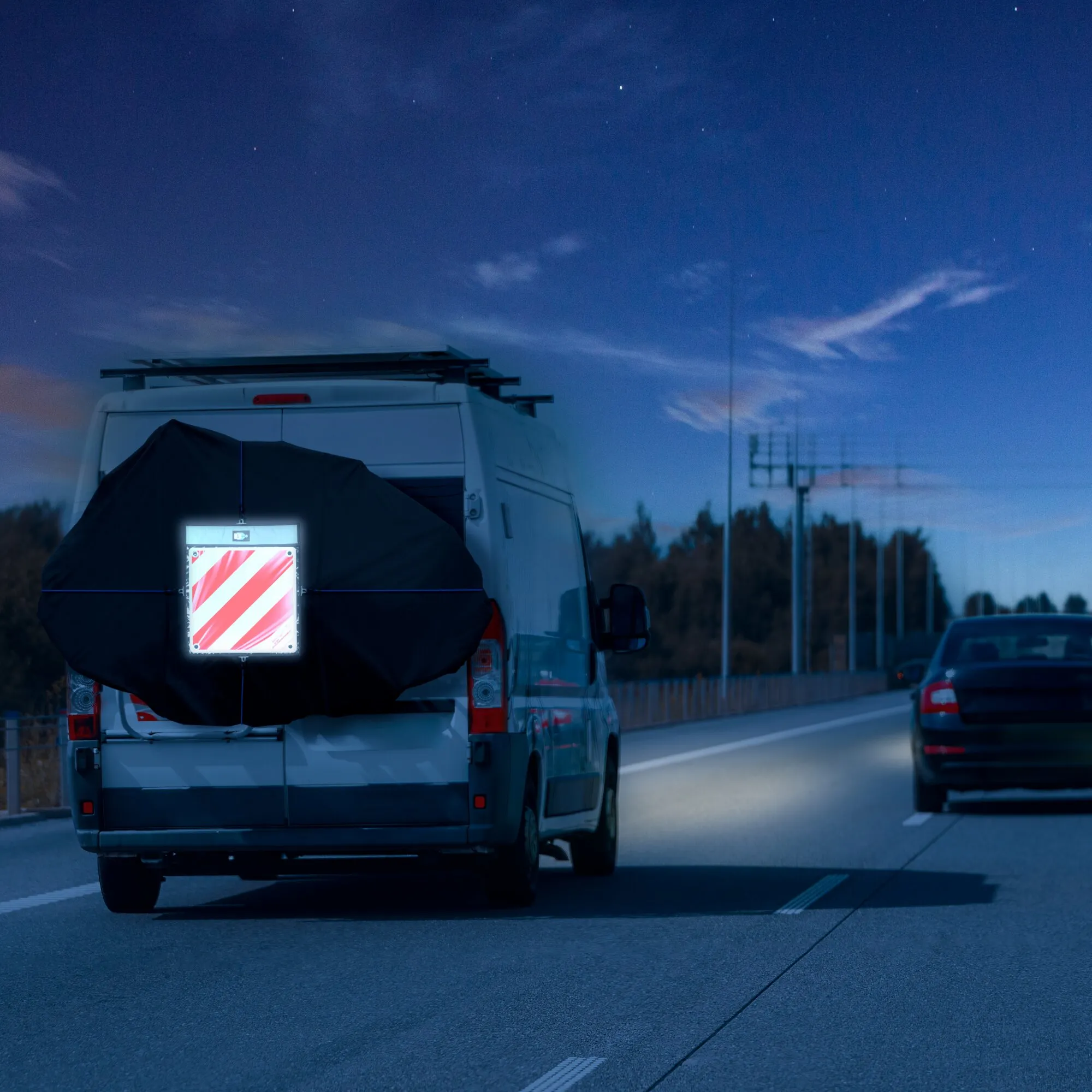 Schwarze Fahrradschutzhülle für zwei bis drei Fahrräder am Heck eines weißen Wohnmobils auf einer nächtlichen Autobahn
