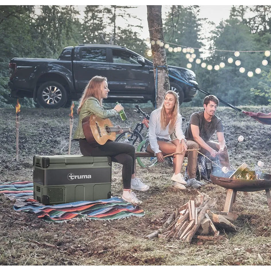 Graue Kompressorkühlbox Truma Cooler mit 72 Litern Fassungsvermögen beim Camping neben drei Personen und Lagerfeuer