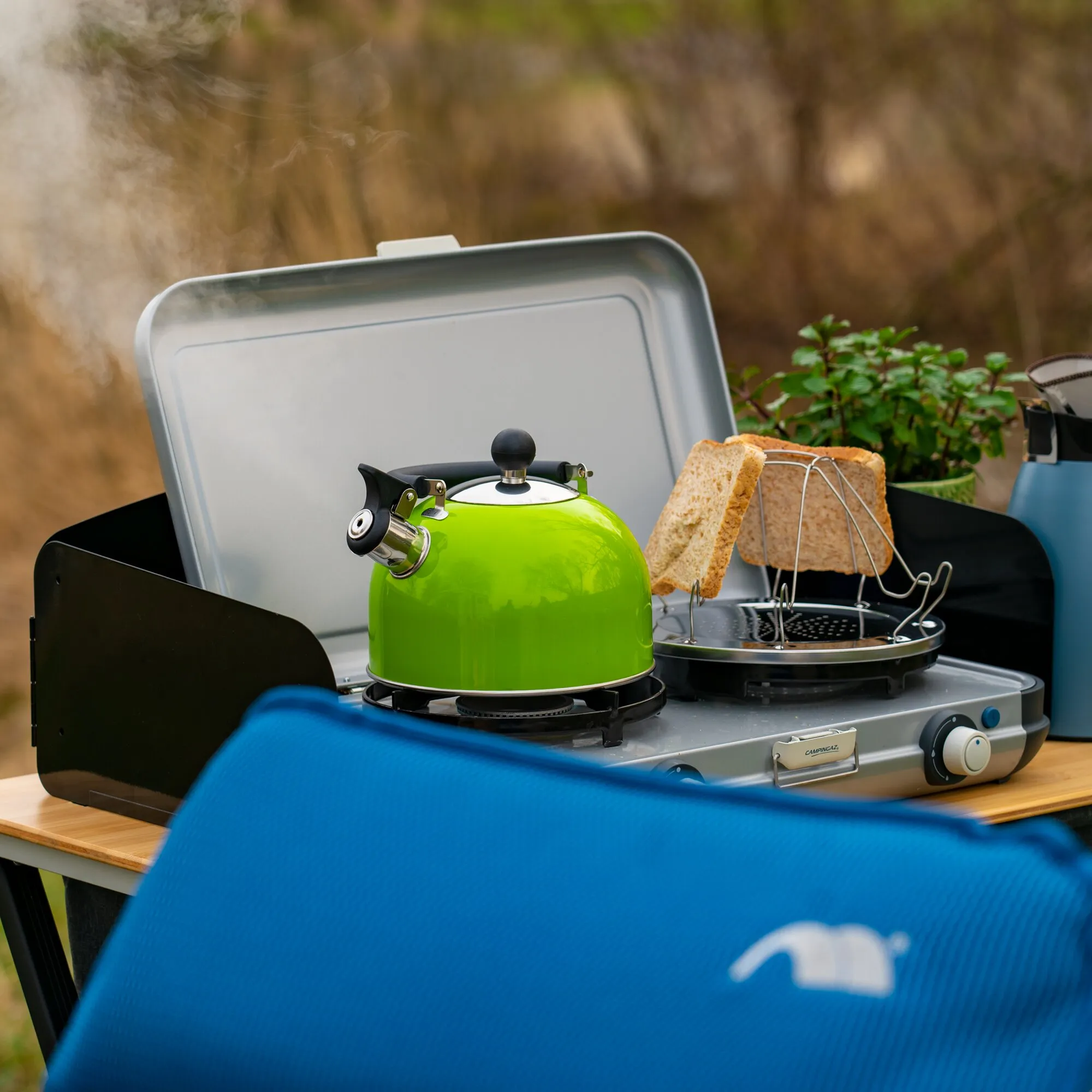 Grüner Flötenkessel mit schwarzem Griff auf einem Campingkocher, daneben getoastetes Brot und aufsteigender Dampf