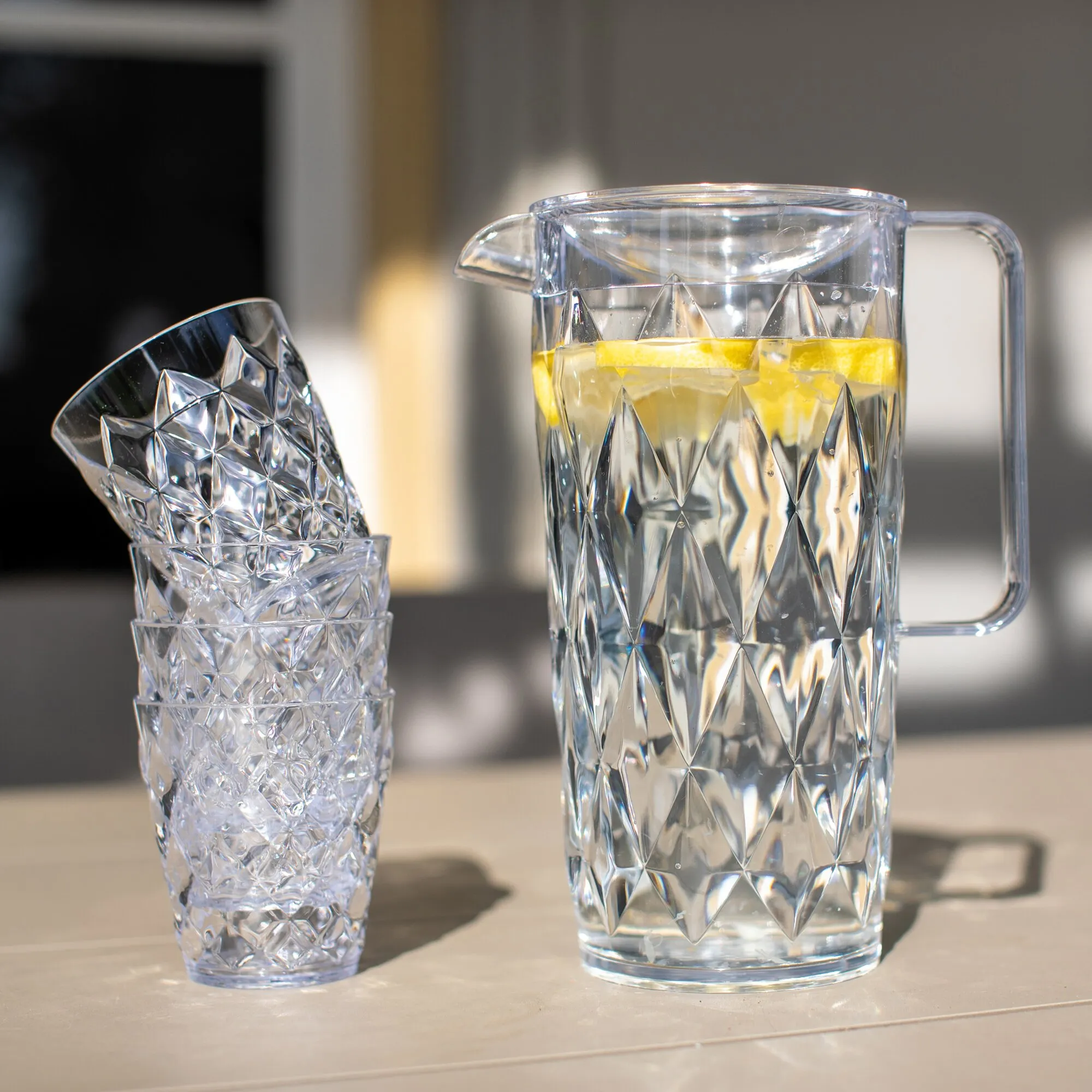 Transparente Kanne CRYSTAL mit geometrischem Schliff, gefüllt mit Zitronenwasser, daneben vier passende strukturierte Becher