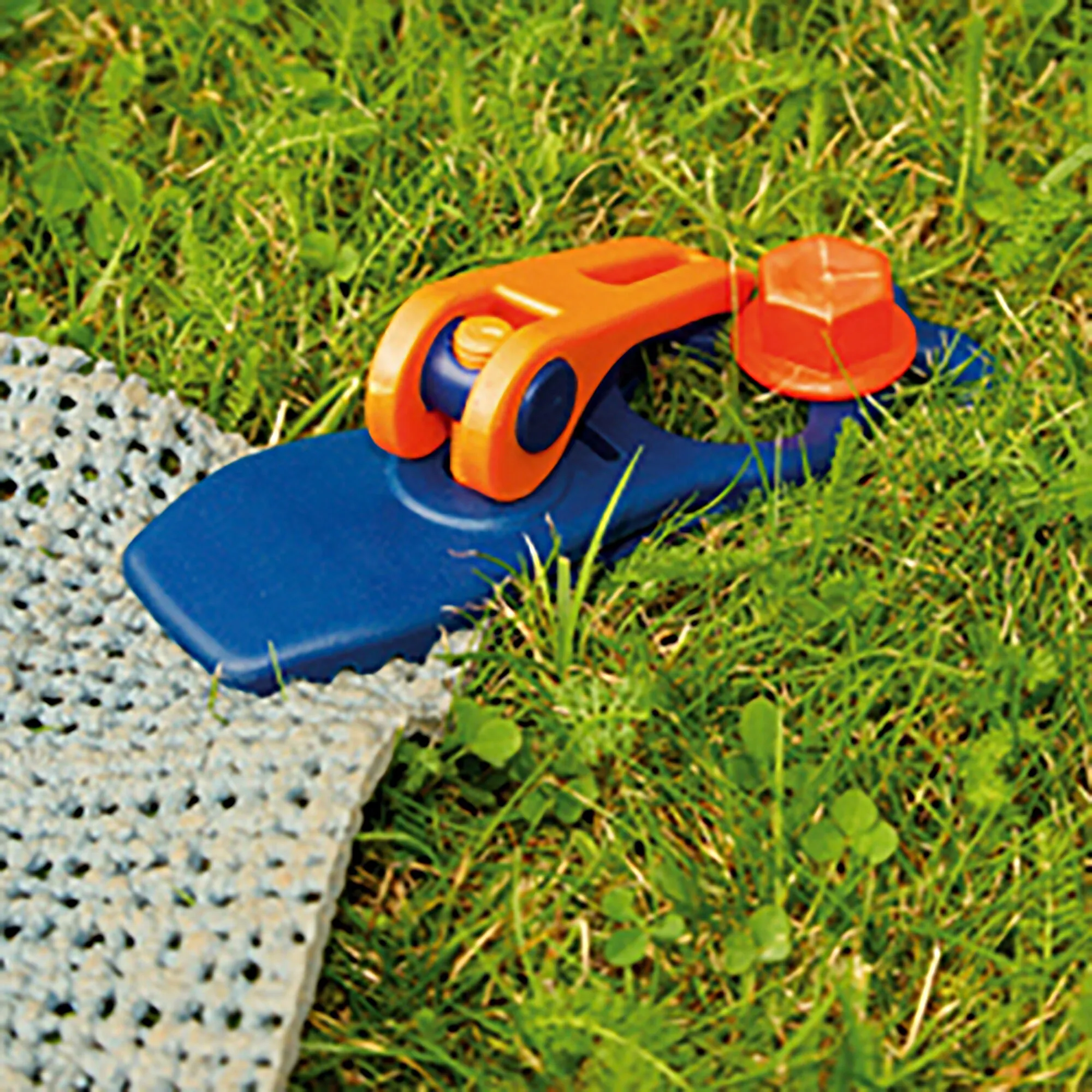 Orange-blaue Schnellspannklemme PP Crocodile mit großem Griff klemmt eine blaue Platte am Rand einer Rasenfläche