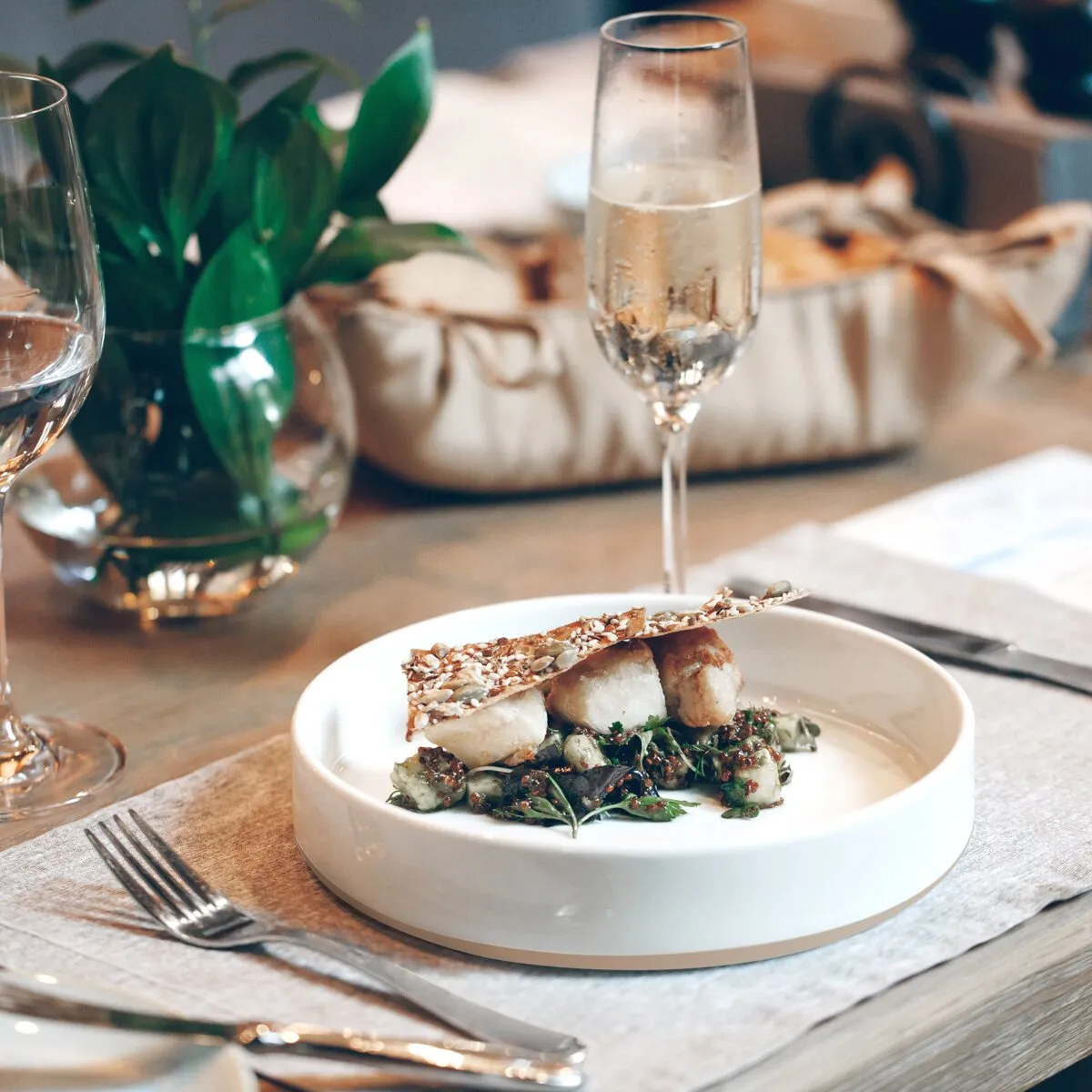 Weißes, rundes Lunch-Set Odette mit angerichtetem Gericht auf einem gedeckten Holztisch neben Weingläsern und Besteck