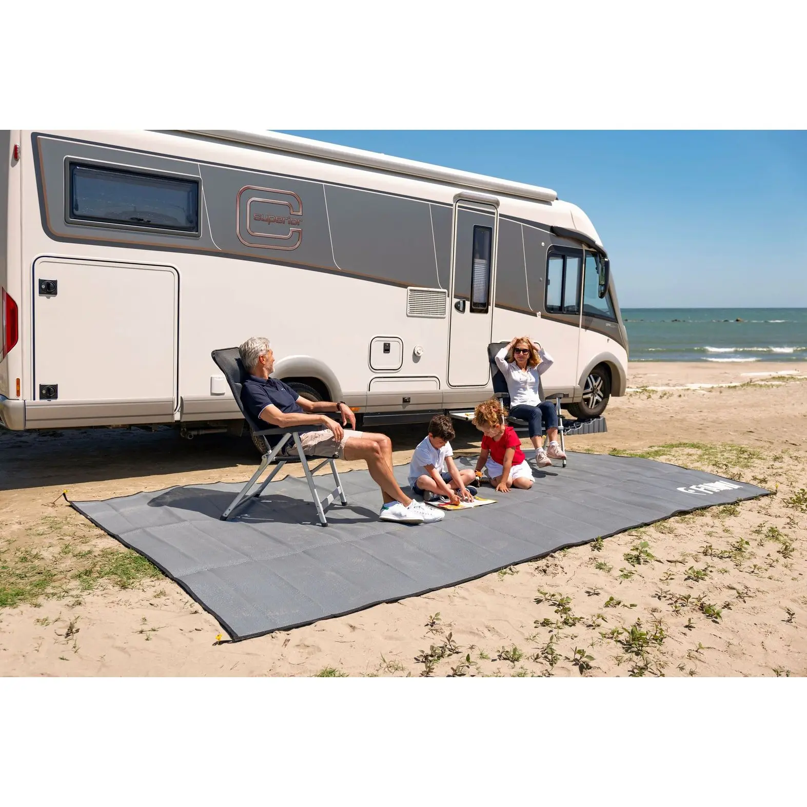 Großer dunkelgrauer Zeltteppich liegt am Strand vor einem Wohnmobil; eine Familie sitzt entspannt auf Stühlen, die Kinder malen auf dem Teppich