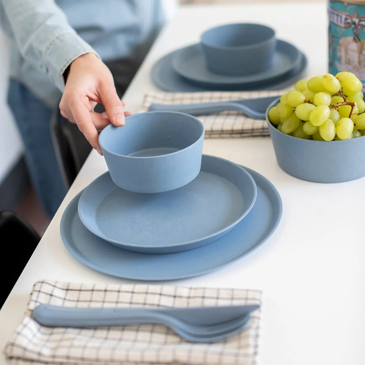 Blaues Besteckset KLIKK mit Löffel und Gabel liegt auf kariertem Tuch neben blauem Geschirr und einer Schale Weintrauben