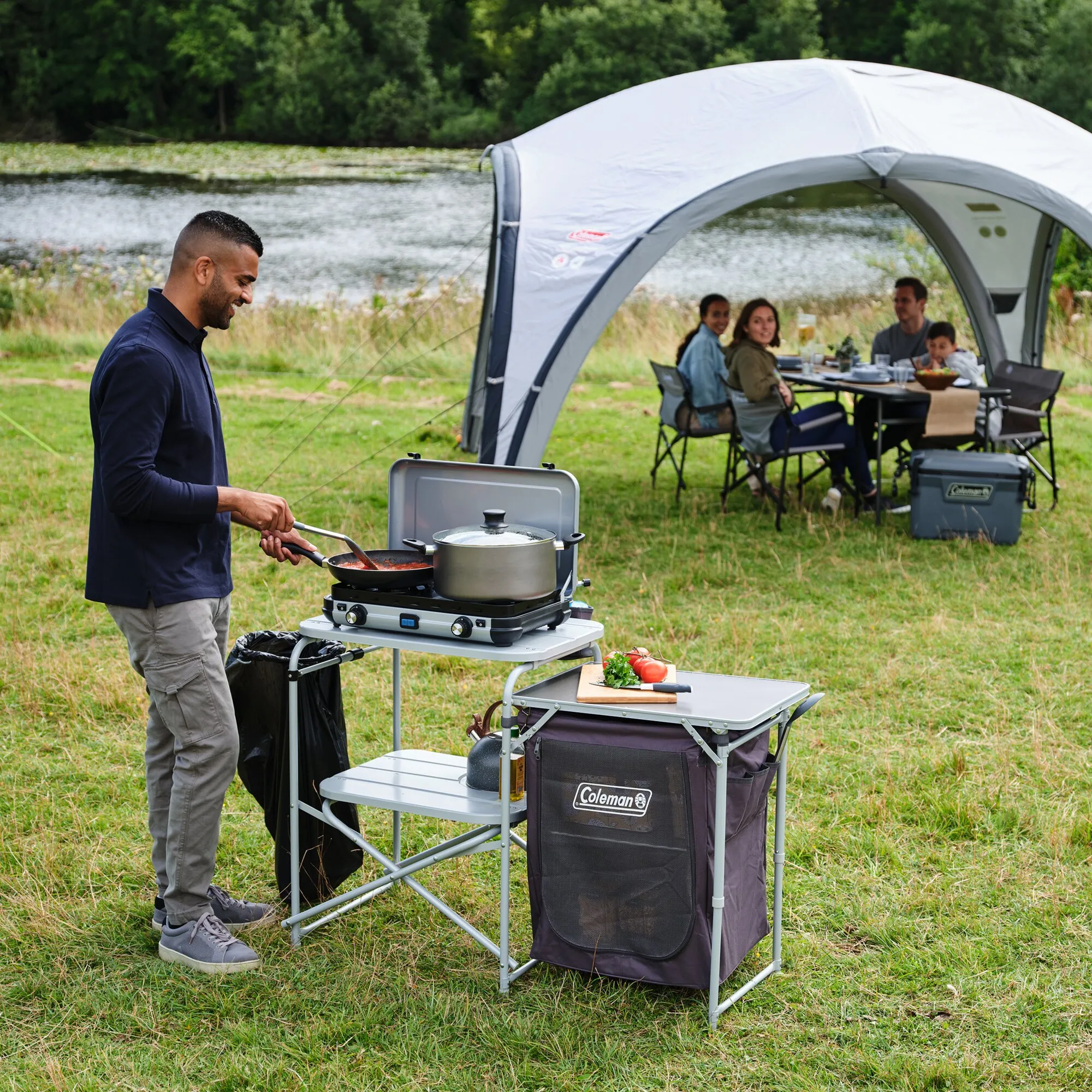 Campingküche Cooking Stand mit zweiflammigem Kocher, Töpfen und Ablage; ein Mann kocht davor im Freien neben einem Zelt