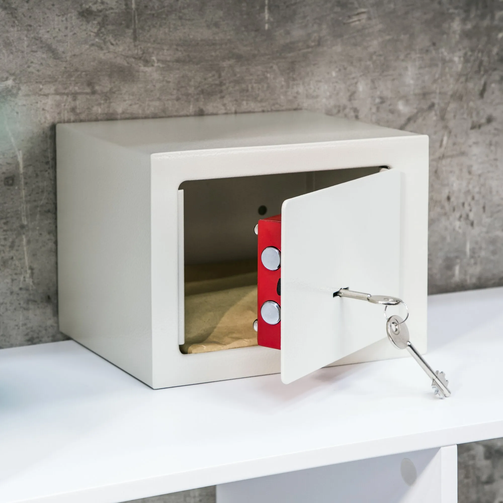 Weißer Mini-Tresor mit geöffnetem Deckel, roter Verriegelung und eingestecktem Schlüssel auf einem weißen Tisch