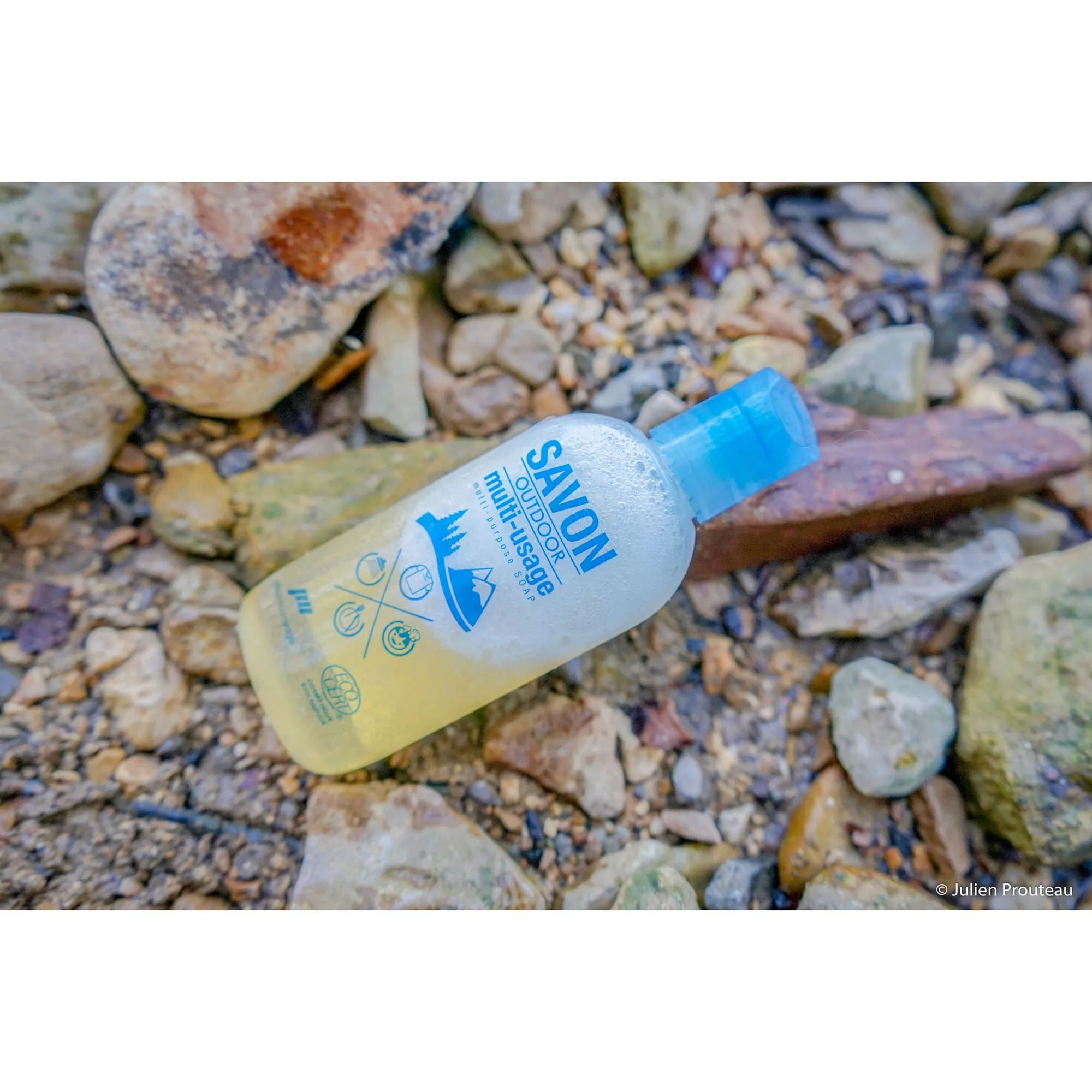 Transparente Flasche mit blauem Verschluss und gelber SAVON OUTDOOR Multi-Usage-Seife auf einem steinigen Bachbett