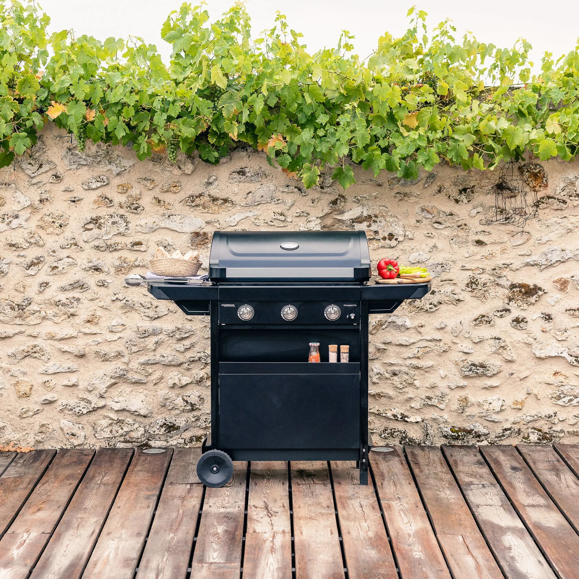 Schwarzer Gasgrill Compact 3 L mit geschlossenem Deckel, drei Bedienknöpfen und seitlichen Ablagen vor einer Natursteinwand 
