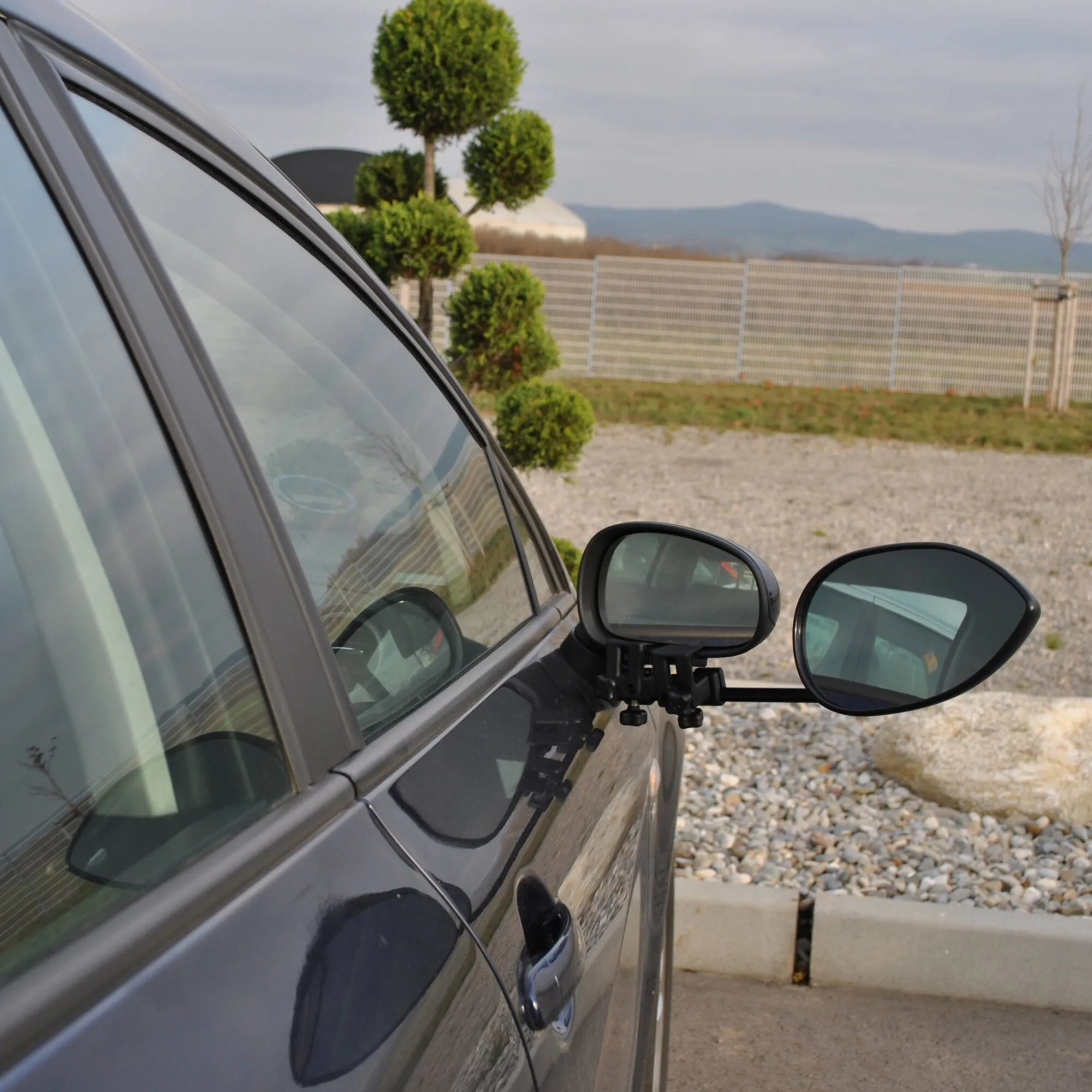 Schwarzes Auto mit montiertem Caravanspiegel Aero 4 Mirror Flat neben dem Außenspiegel, aufgenommen in einer Landschaft mit Bergen