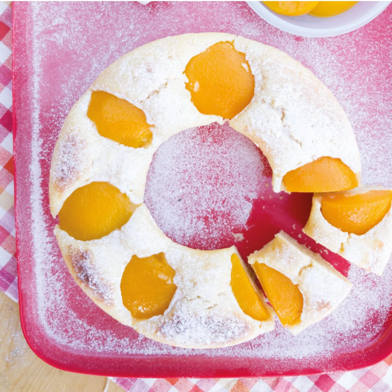 Runder, mit Puderzucker bestäubter Obstkuchen mit Aprikosenstücken und einer großen Pflaume in der Mitte auf rotem Brett