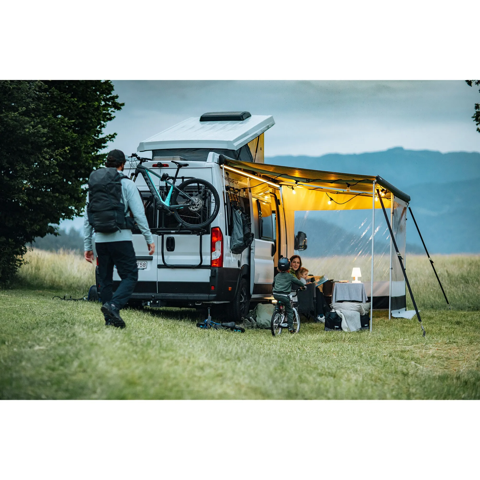 Ausgefahrene Thule Omnistor 5200 Markise an einem weißen Campervan, darunter sitzt eine Familie beim Camping auf einer Wiese