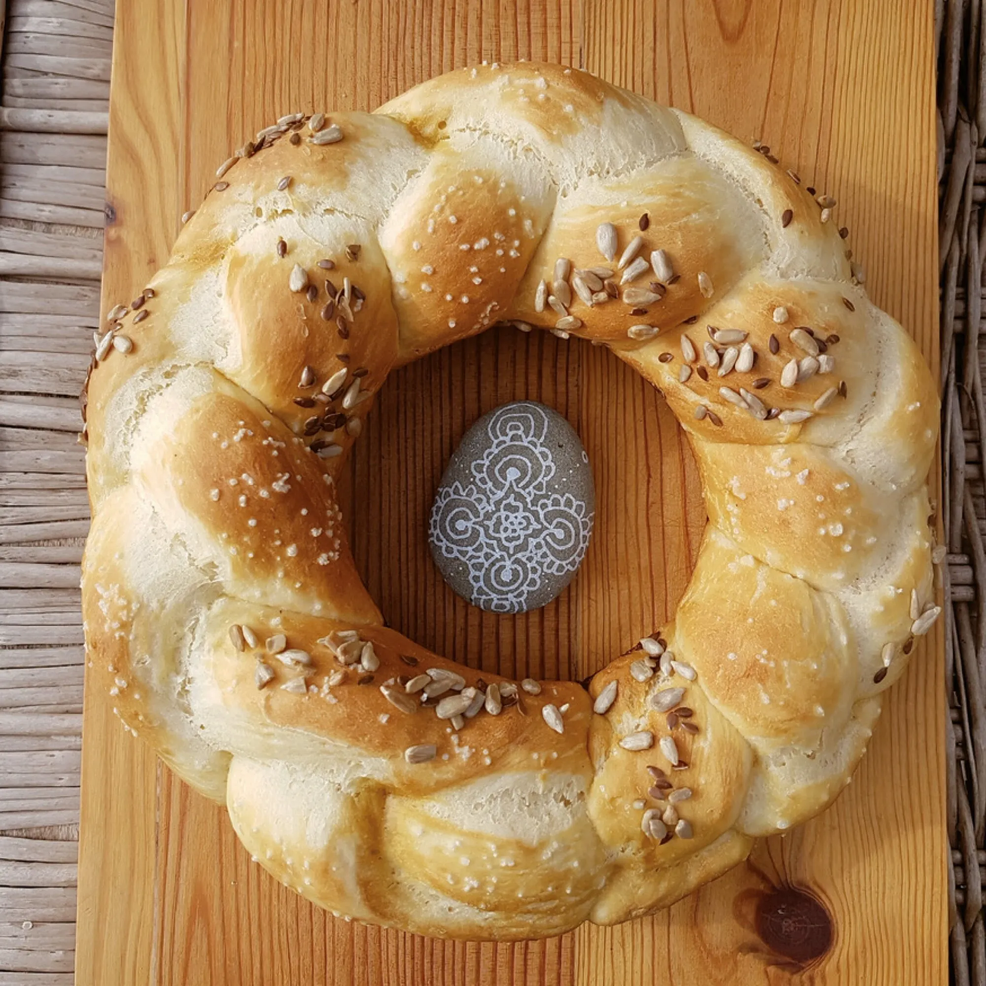 Goldbraunes, geflochtenes Ringbrot mit Sonnenblumenkernen und Salz auf einem Holzbrett, in der Mitte ein verziertes Ei