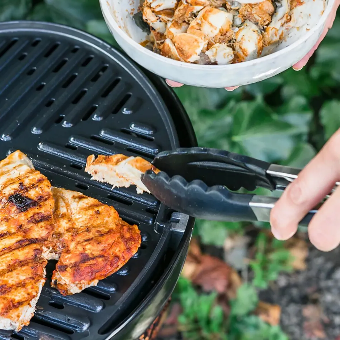 Edelstahl-Grillzange mit schwarzen Greifbacken beim Wenden eines Hähnchenstücks auf dem Grill, Schüssel mit mariniertem Fleisch im Hintergrund