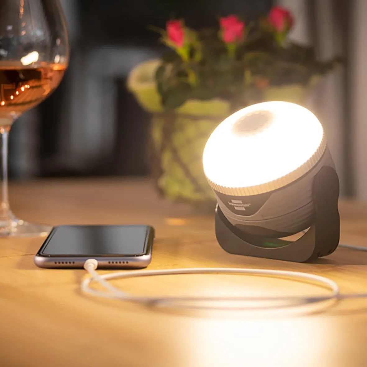 Leuchtende schwarze Campingleuchte OLI 310 AB mit Smartphone und Ladekabel auf einem Holztisch, Blumen im Hintergrund
