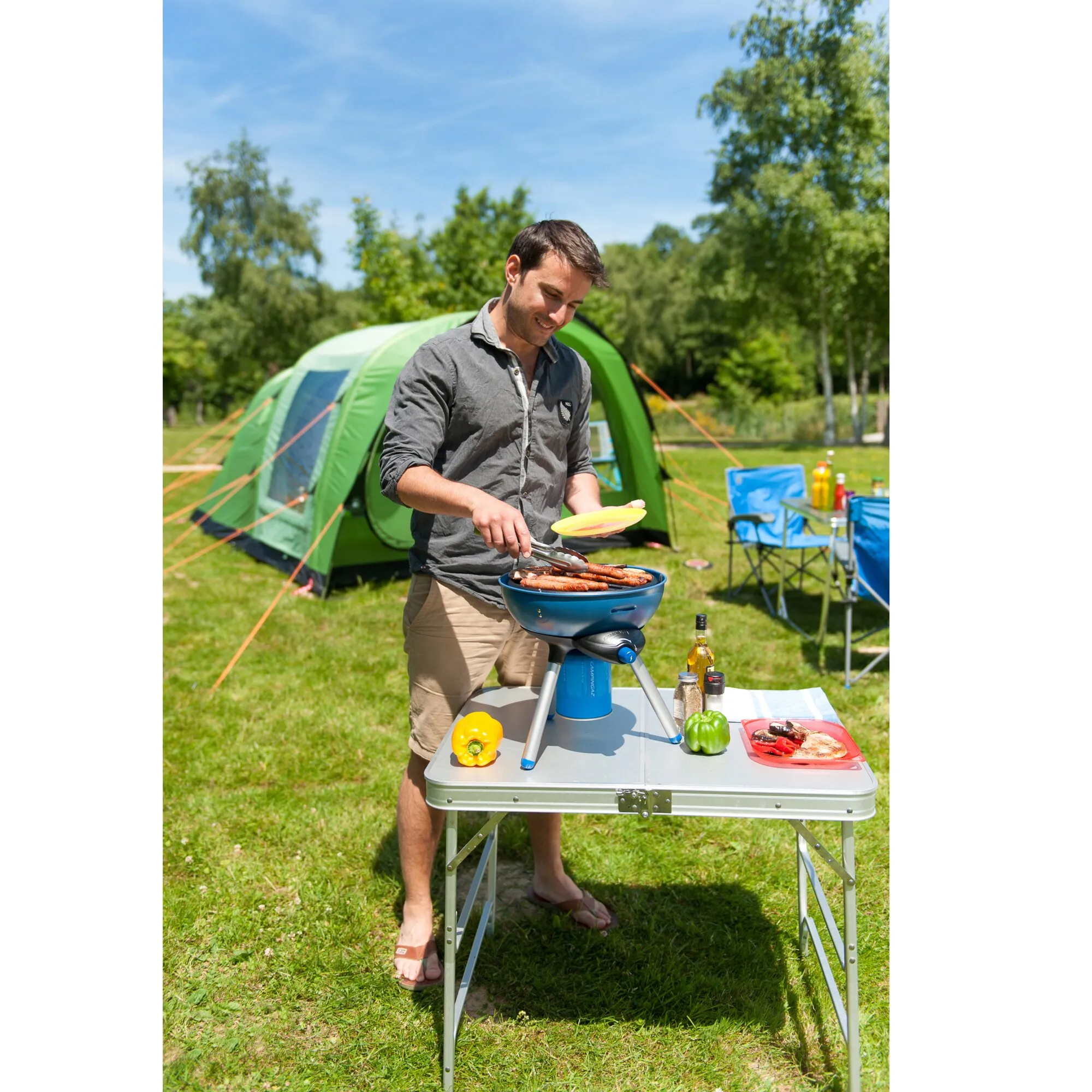 Mann grillt Würstchen auf dem blauen Campinggrill Party Grill 200 CV vor einem Zelt auf einer Wiese