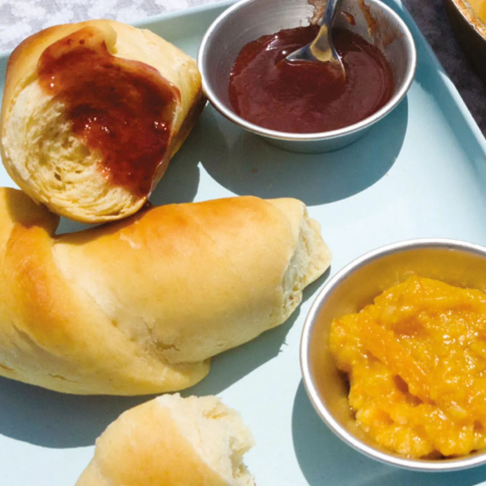 Aufgeschnittene, goldbraune Brötchen mit roter und orangefarbener Marmelade in Metallschalen, passend zum Brotbackbuch für den Omnia Backofen