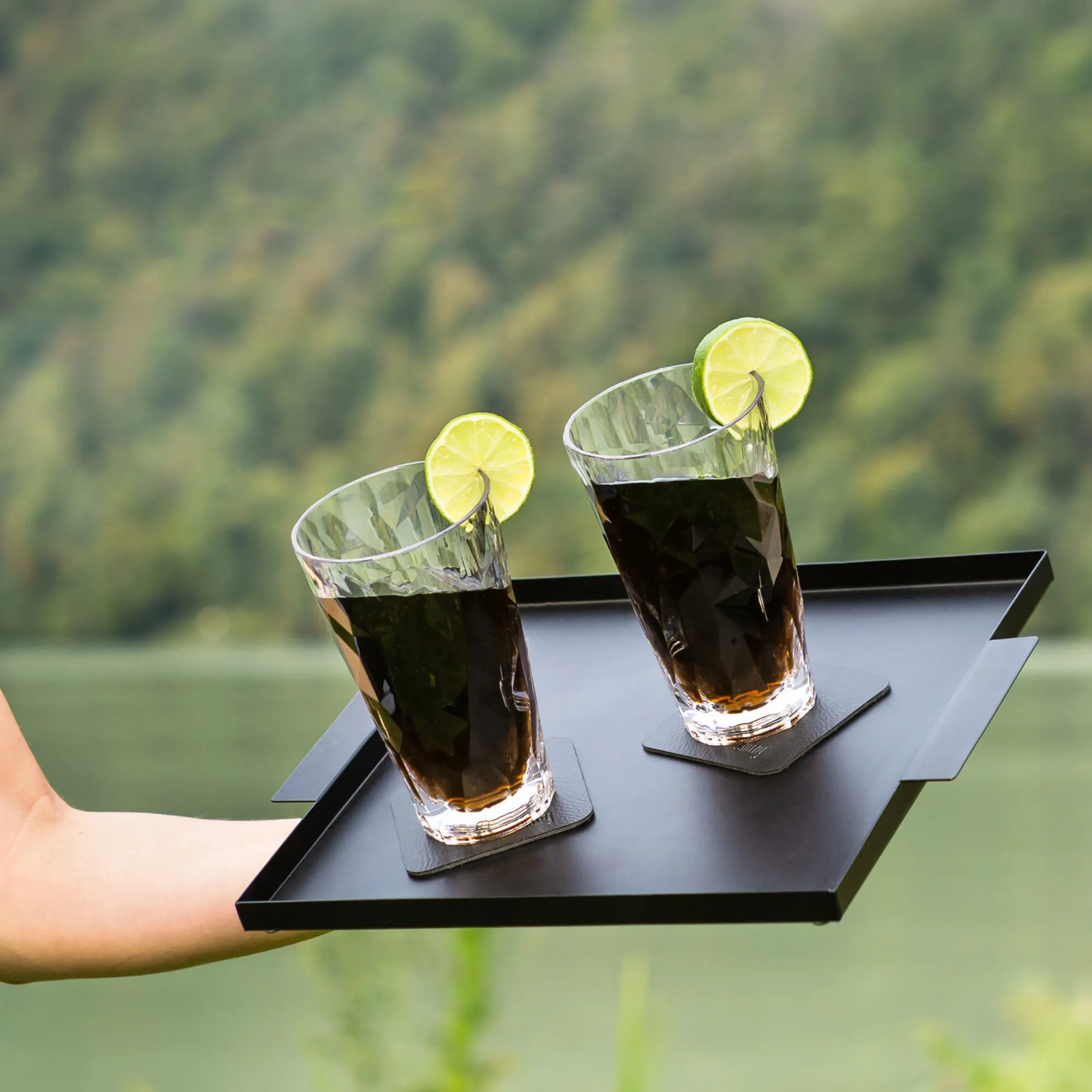 Zwei transparente Longdrink-Kunststoffgläser mit dunklen Getränken und Limettenscheiben auf schwarzen Untersetzern auf einem Tablett