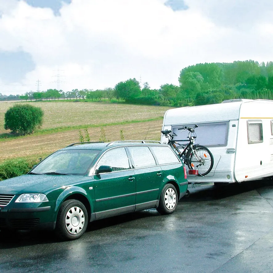 Grüner Kombi mit Wohnwagen und zwei Fahrrädern auf einem Fahrradträger an der Deichsel vor ländlicher Landschaft