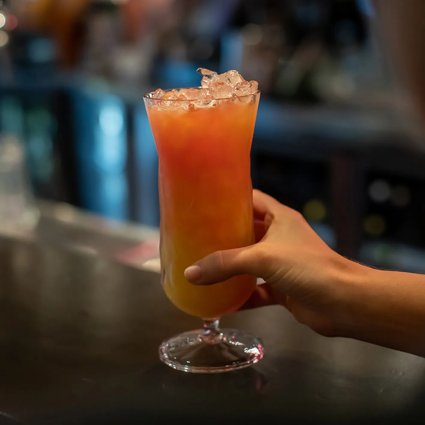 Cocktailglas Superglas CLUB mit orangefarbenem Drink, Eiswürfeln und Stiel, von einer Hand an einer Bar gehalten