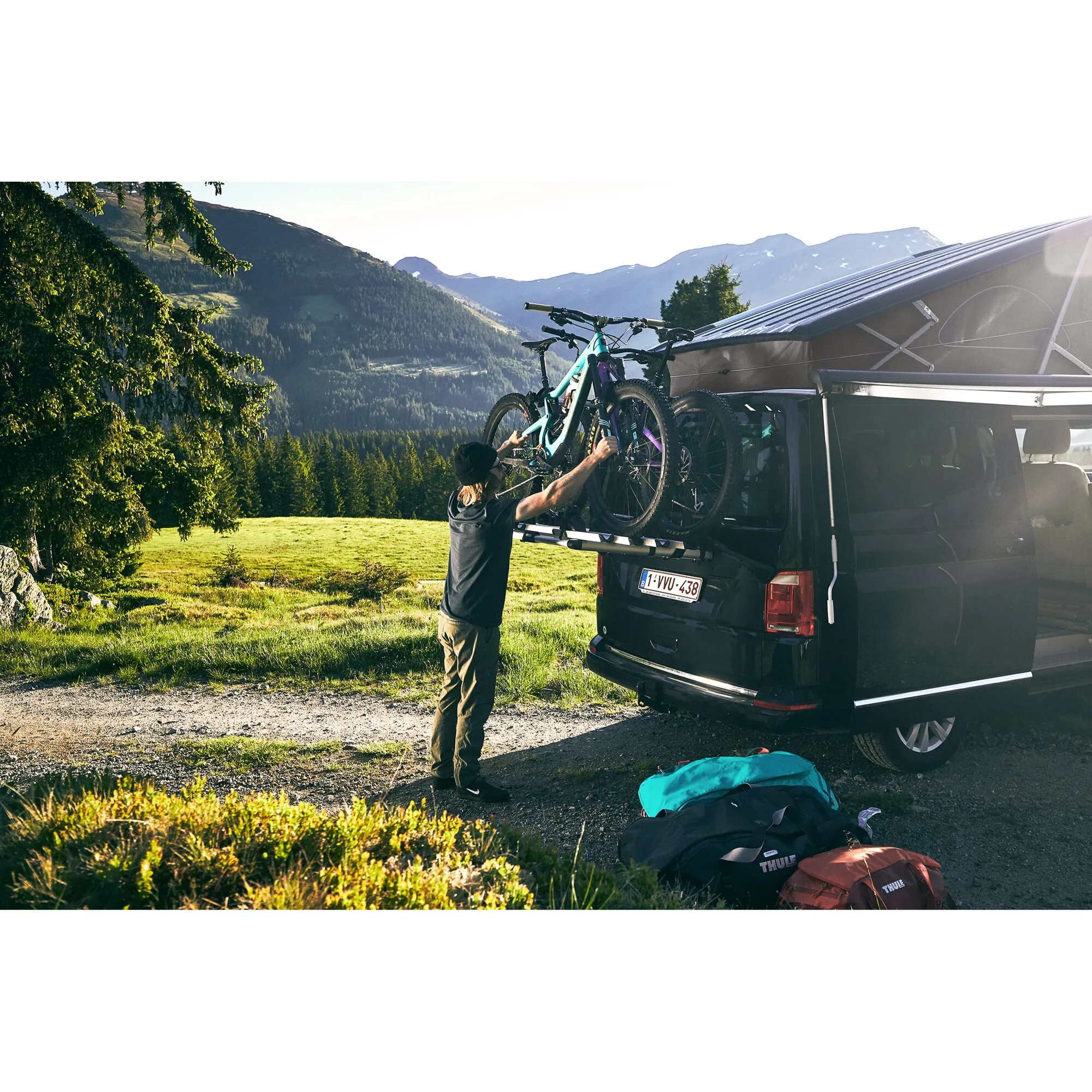 Schwarzer VW T6 mit Thule WanderWay Fahrradträger und zwei Mountainbikes vor alpiner Berglandschaft, eine Person befestigt ein Fahrrad