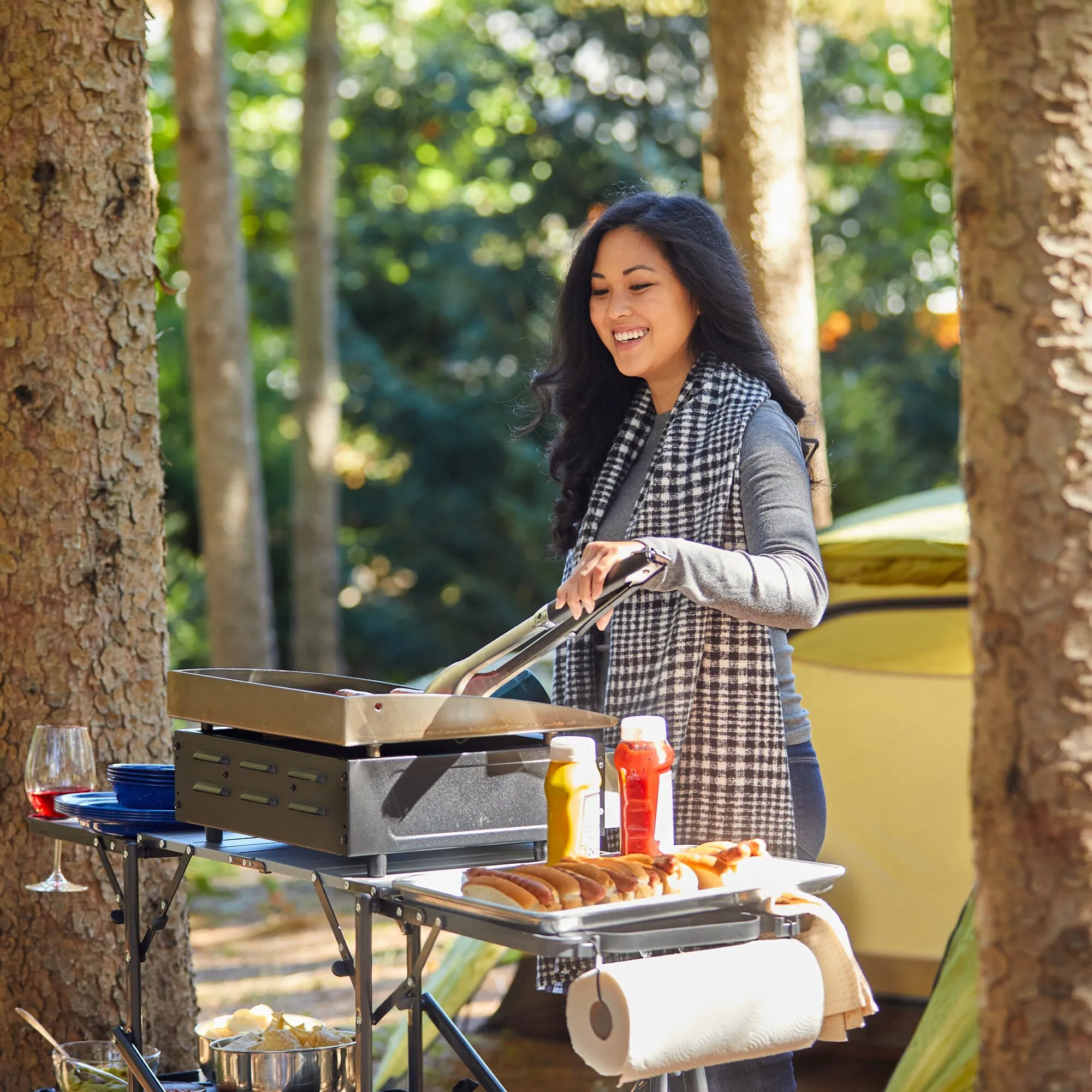 Frau bereitet im Wald an einer mobilen schwarzen Campingküche mit Grillplatte Würstchen und Beilagen zu