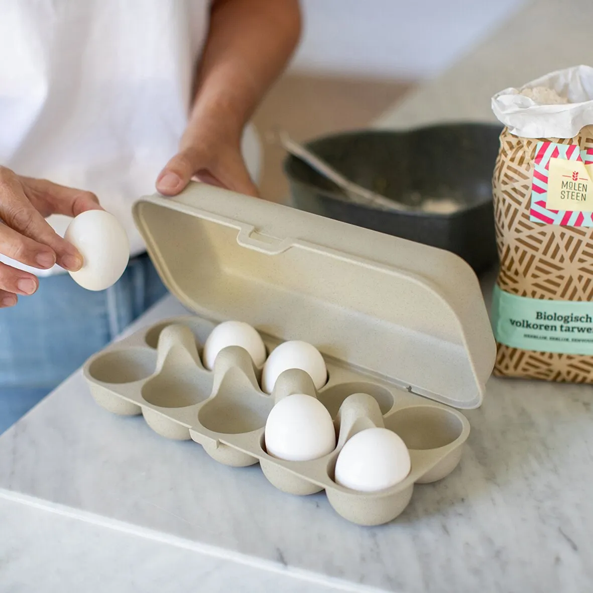 Sandfarbene Eierbox EGGS TO GO mit geöffnetem Deckel und vier weißen Eiern, während ein weiteres hineingelegt wird