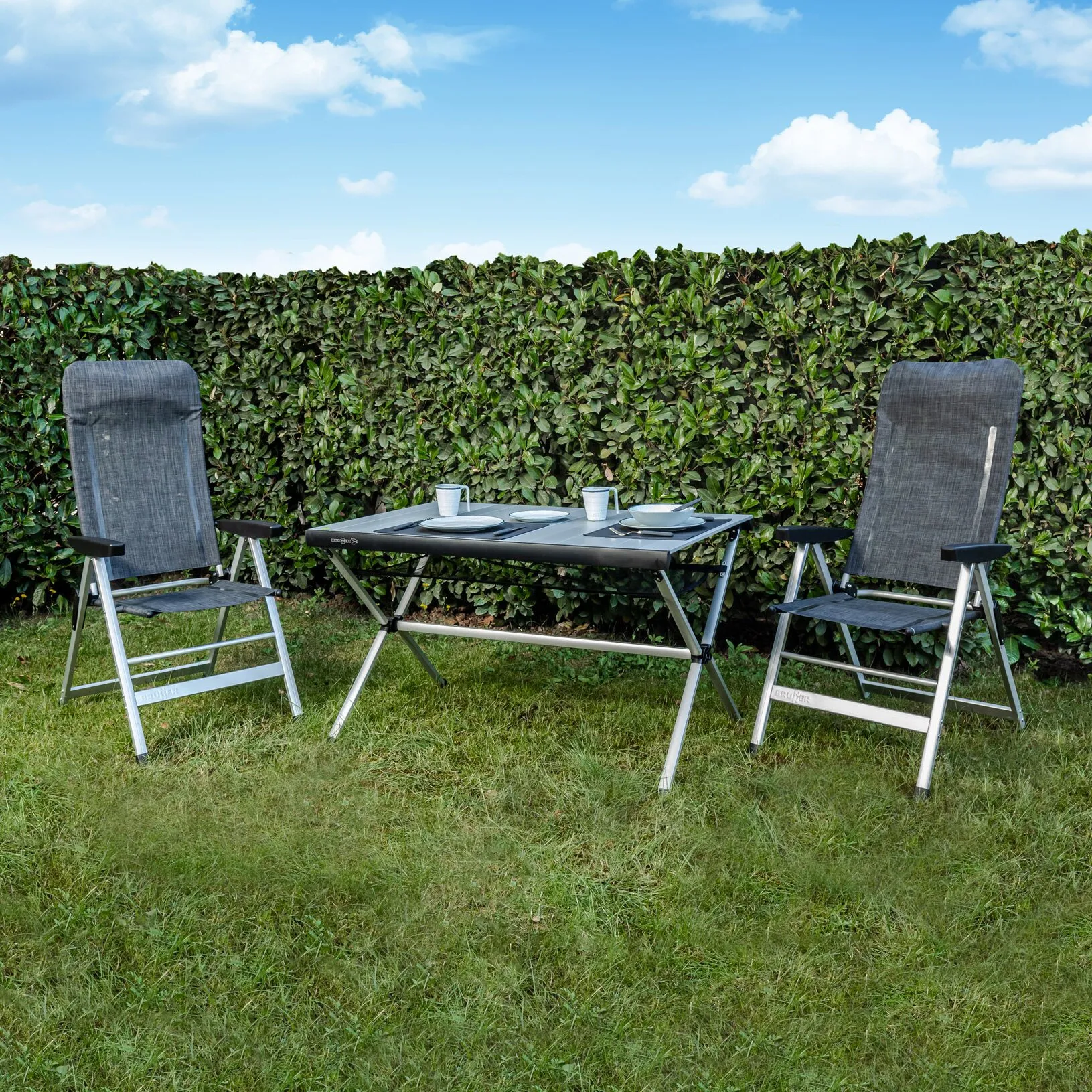Zwei graue Campingstühle Skye Syntex Plus mit Armlehnen neben einem gedeckten Campingtisch auf einer Wiese vor einer Hecke