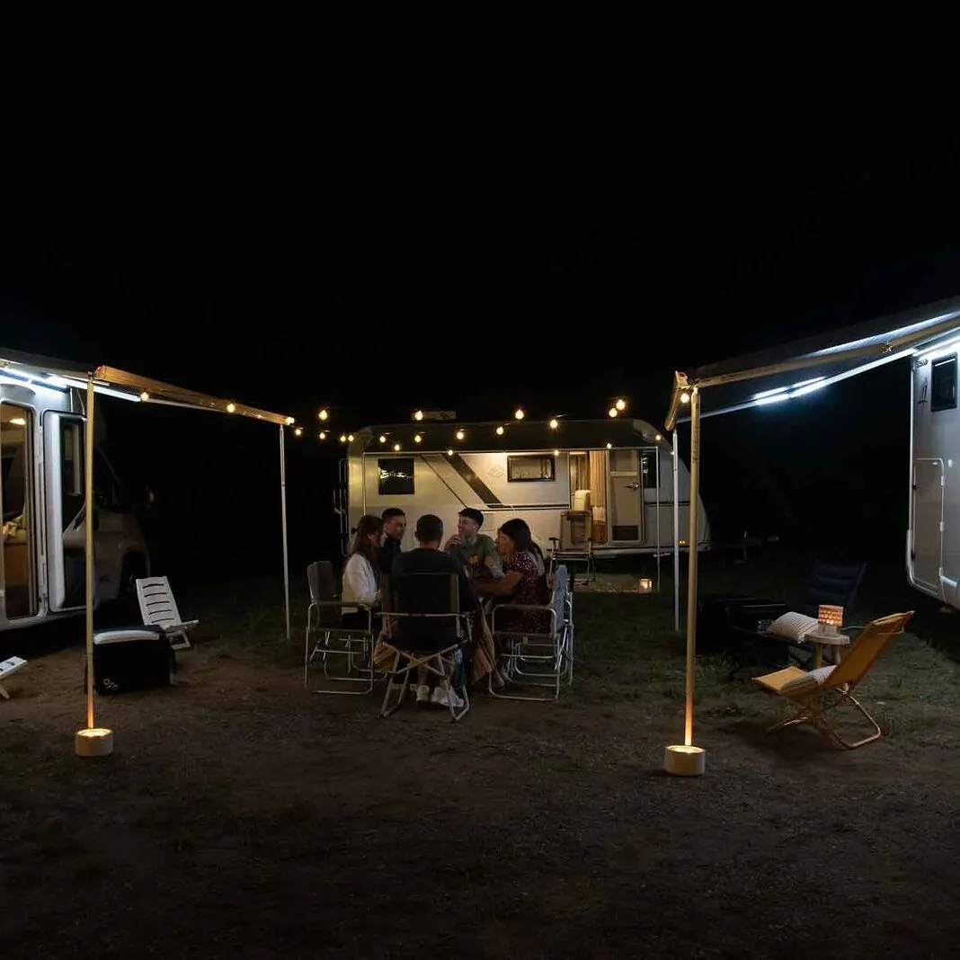 Nächtliche Camping-Szene mit Wohnwagen, ausgefahrenen Markisen und beleuchteten Rafter-LED-Spannstangen, davor sitzende Camper