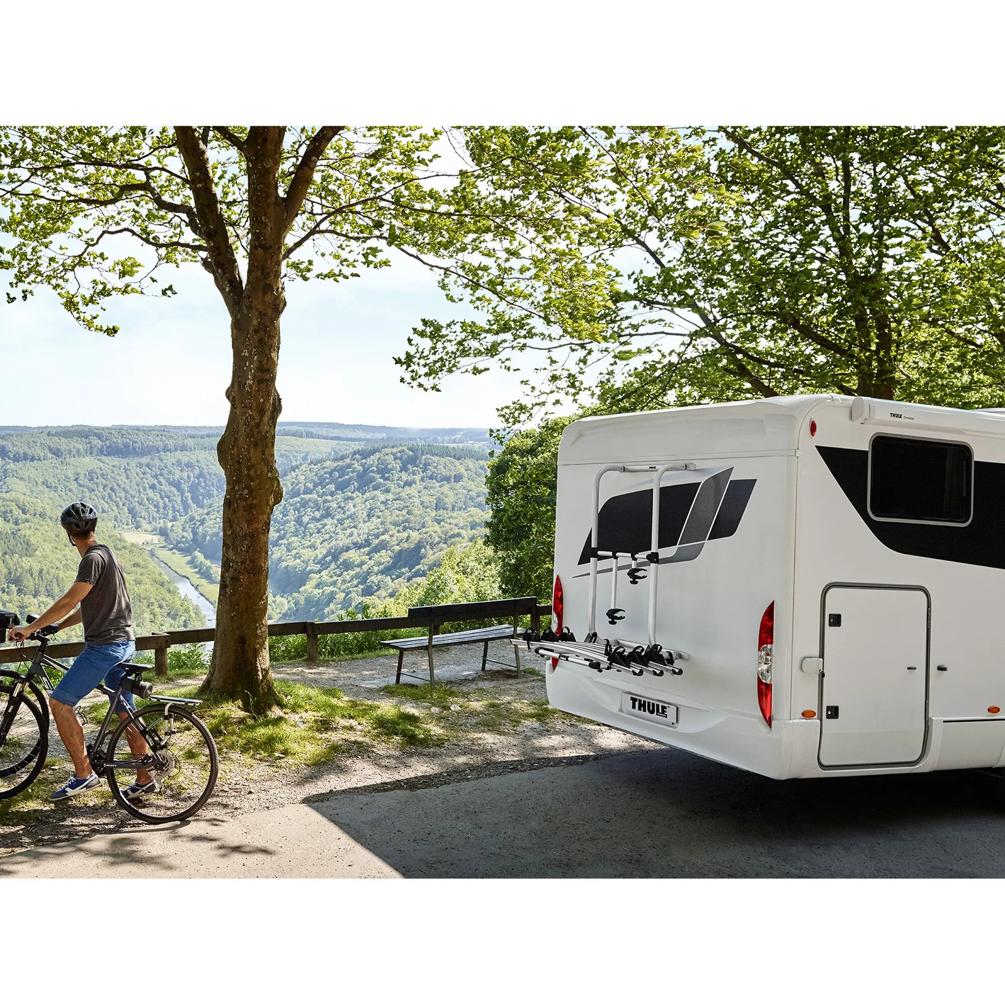 Weißer Wohnwagen mit montiertem Thule Elite G2 HH Fahrradträger am Heck, daneben ein Radfahrer vor bewaldeter Flusslandschaft