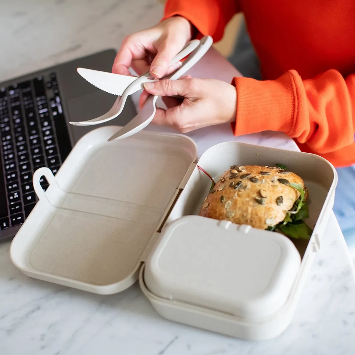 Sandfarbenes Besteckset KLIKK mit Messer, Gabel und Löffel über einer geöffneten Lunchbox mit belegtem Brötchen