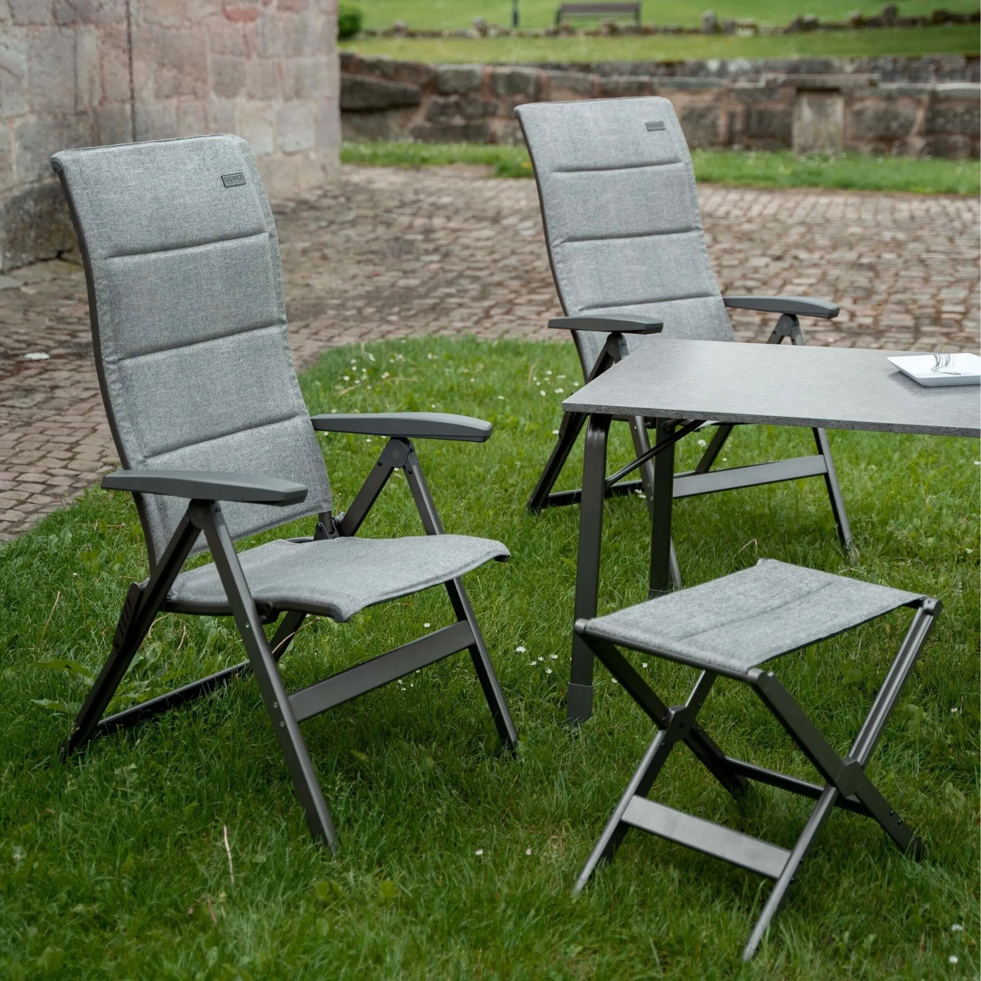 Zwei graue Campingstühle Elegance SG mit Armlehnen neben einem Klapptisch und Hockern auf einer Rasenfläche im Freien