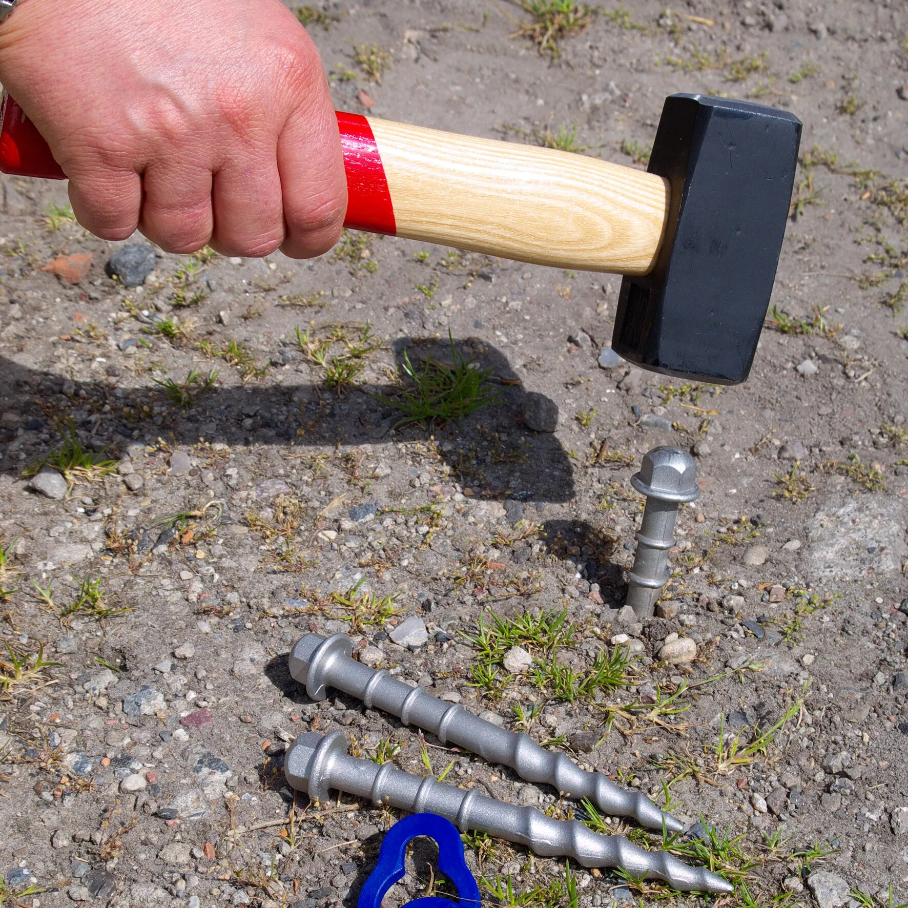 Hand hält einen Vorschlaghammer über einem silbernen PP-Schraubhering im Boden; weitere Schraubheringe liegen daneben