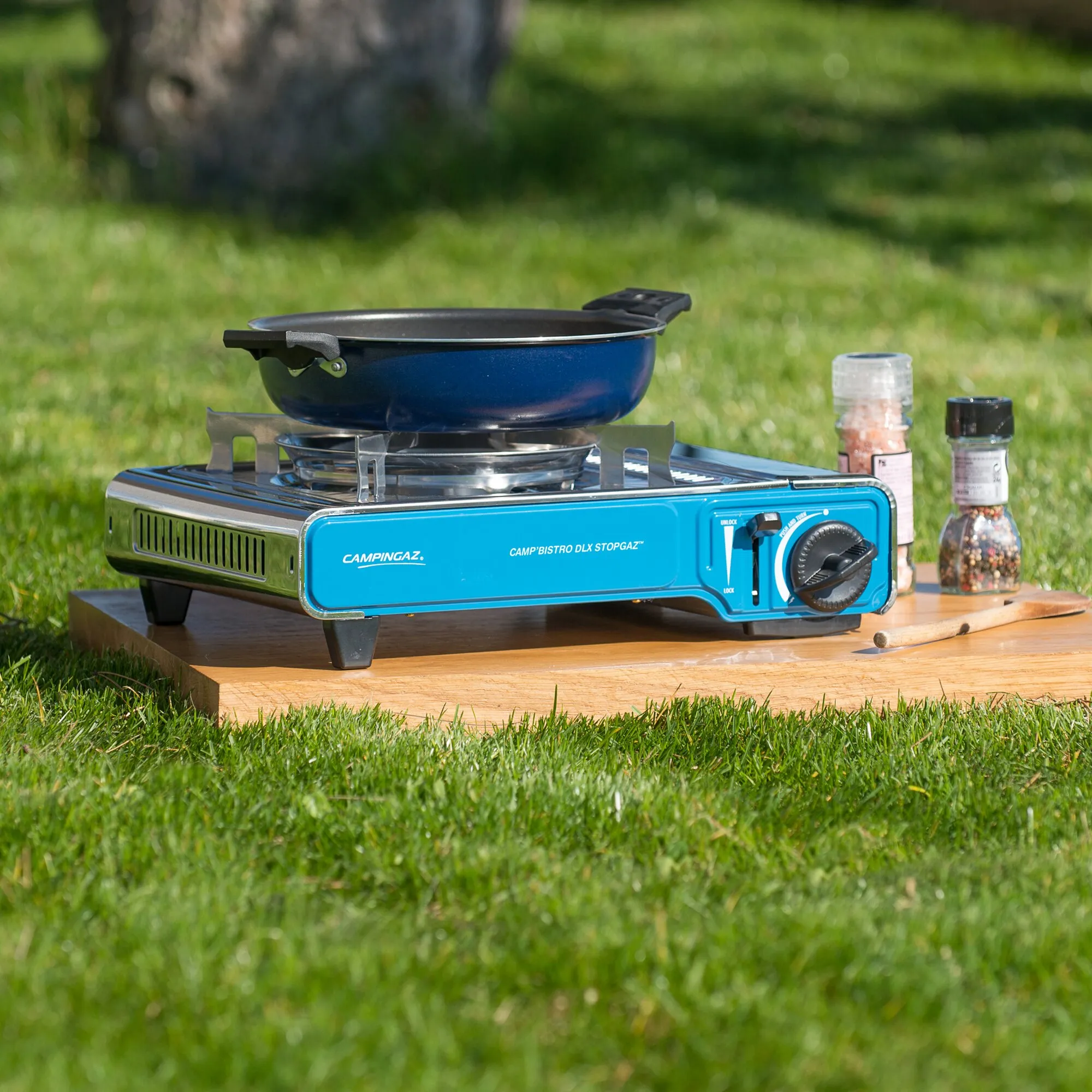 Blauer Campingaz Camp’Bistro DLX Stopgaz Campingkocher mit schwarzer Pfanne auf einem Holztisch im Garten neben Gewürzstreuern
