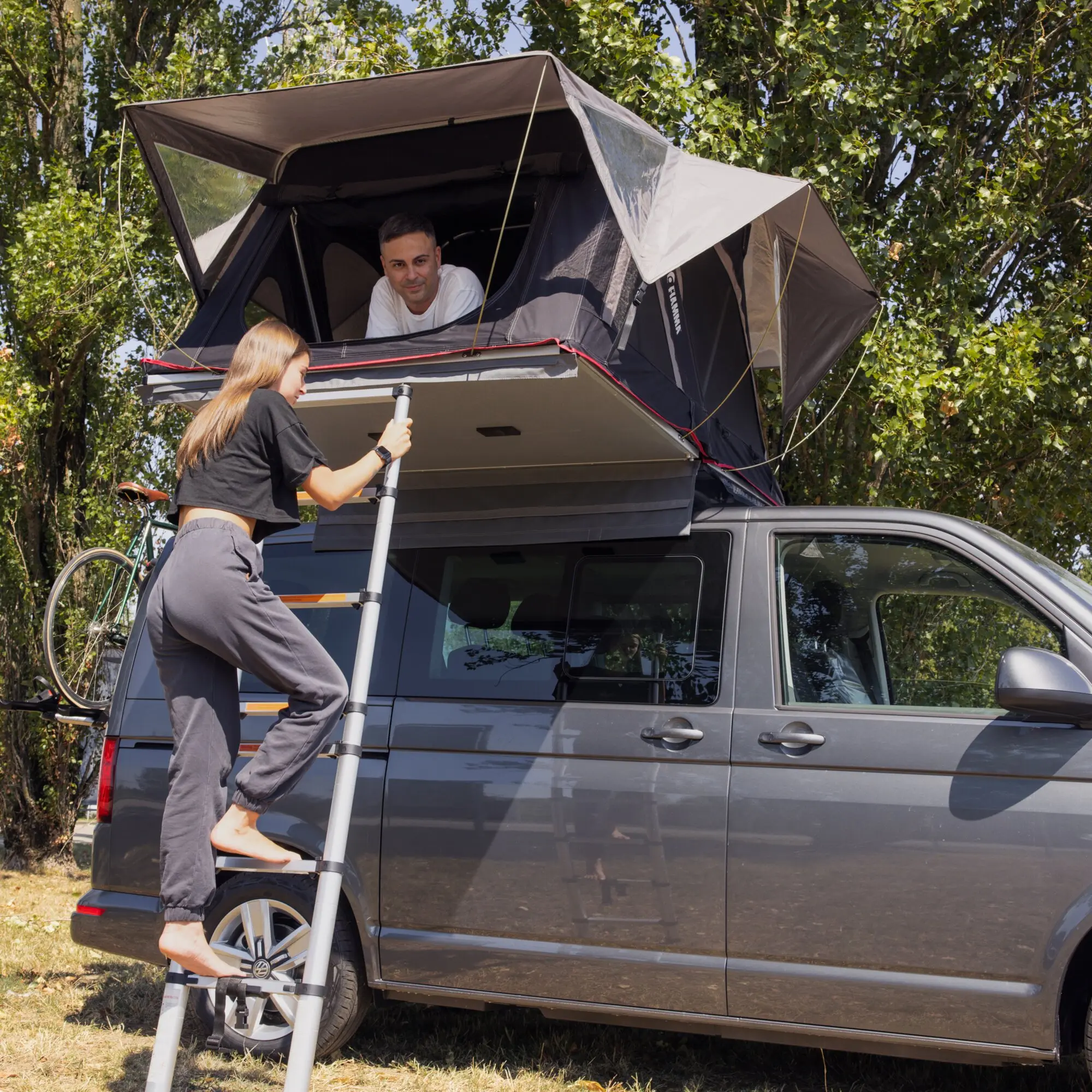 Moonlight Tent 180 cm Dachzelt offen auf Vans Dach, Mann schaut aus Öffnung, Frau auf Leiter daneben, Bäume im Hintergrund