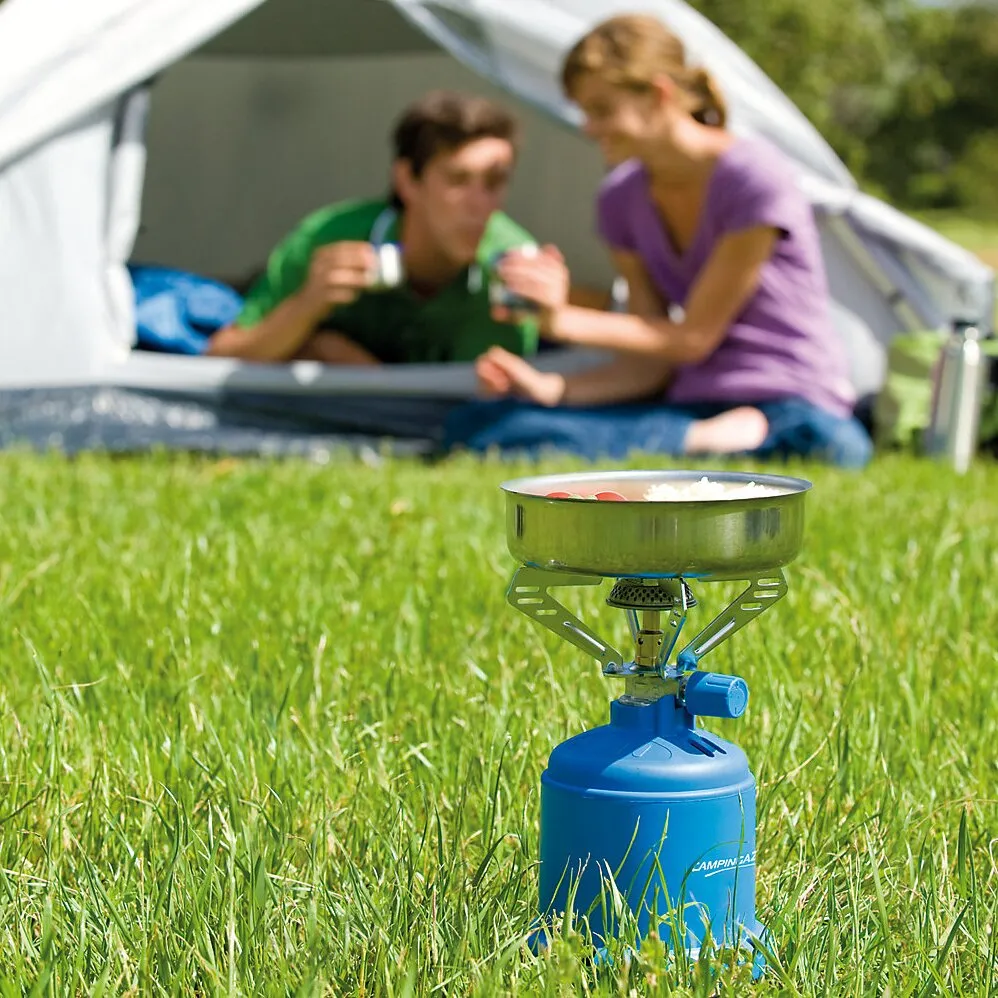 Blauer Campingkocher 206 S mit Metalltopf auf einer Wiese vor einem Zelt, zwei Personen im Hintergrund unerquicklich