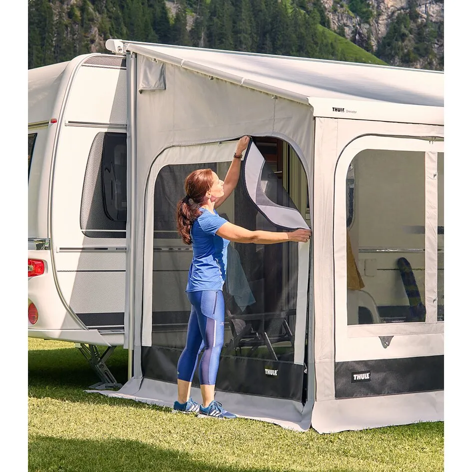 Frau öffnet das Moskitogitter an der Seite eines Thule-Panorama-Vorzelts neben einem Wohnwagen grasbewachsener Campingplatz