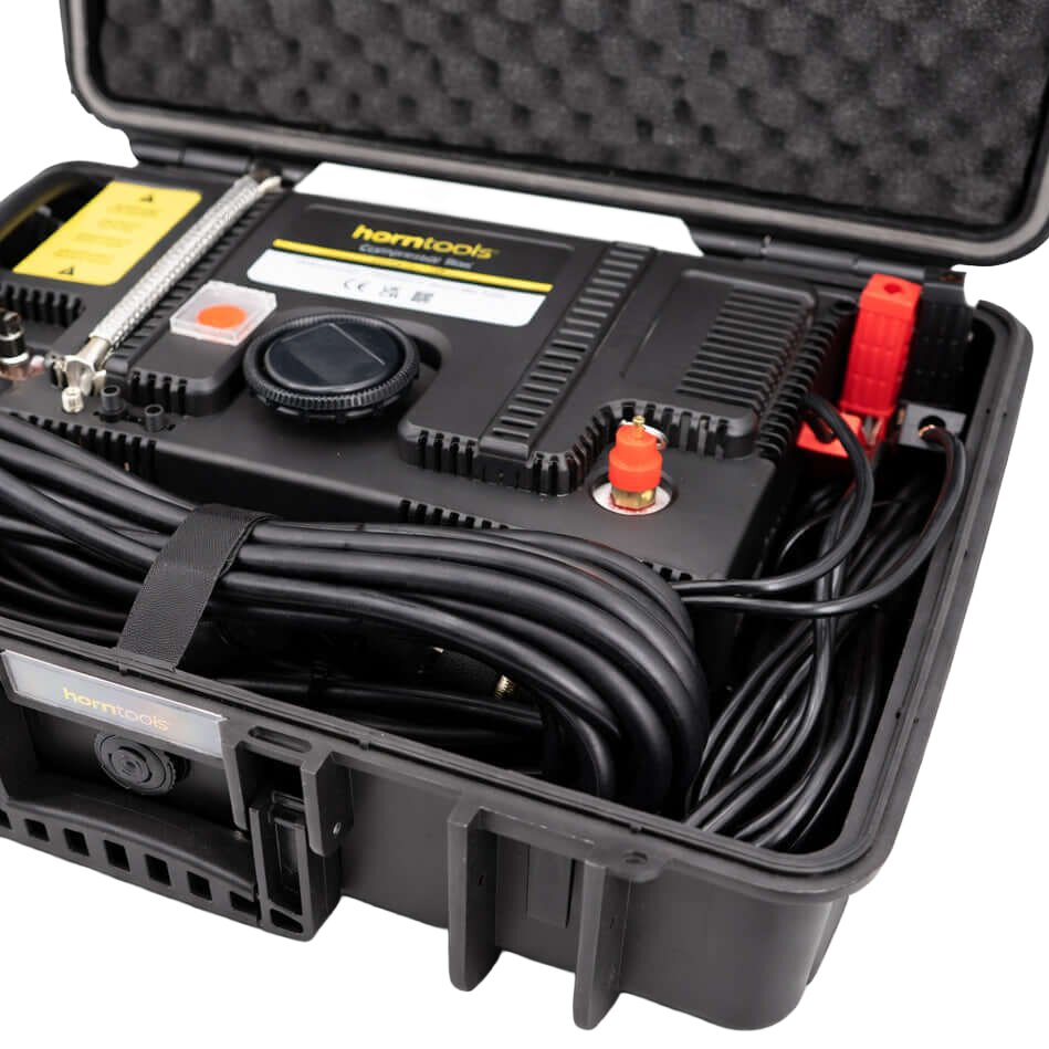 Geöffnete schwarze Kofferbox mit einer kompakten Kompressor-Box, aufgewickeltem Kabel, rundem Einstellrad, Schlauch und roten Klemmen am Rand