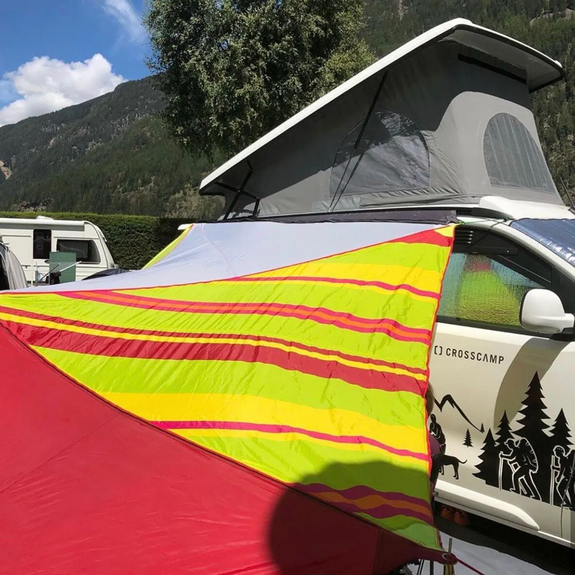 Crosscamp-Camper mit aufgestelltem Dachzelt und bunt gestreiftem Vorzelt auf einem Campingplatz vor bewaldeten Bergen