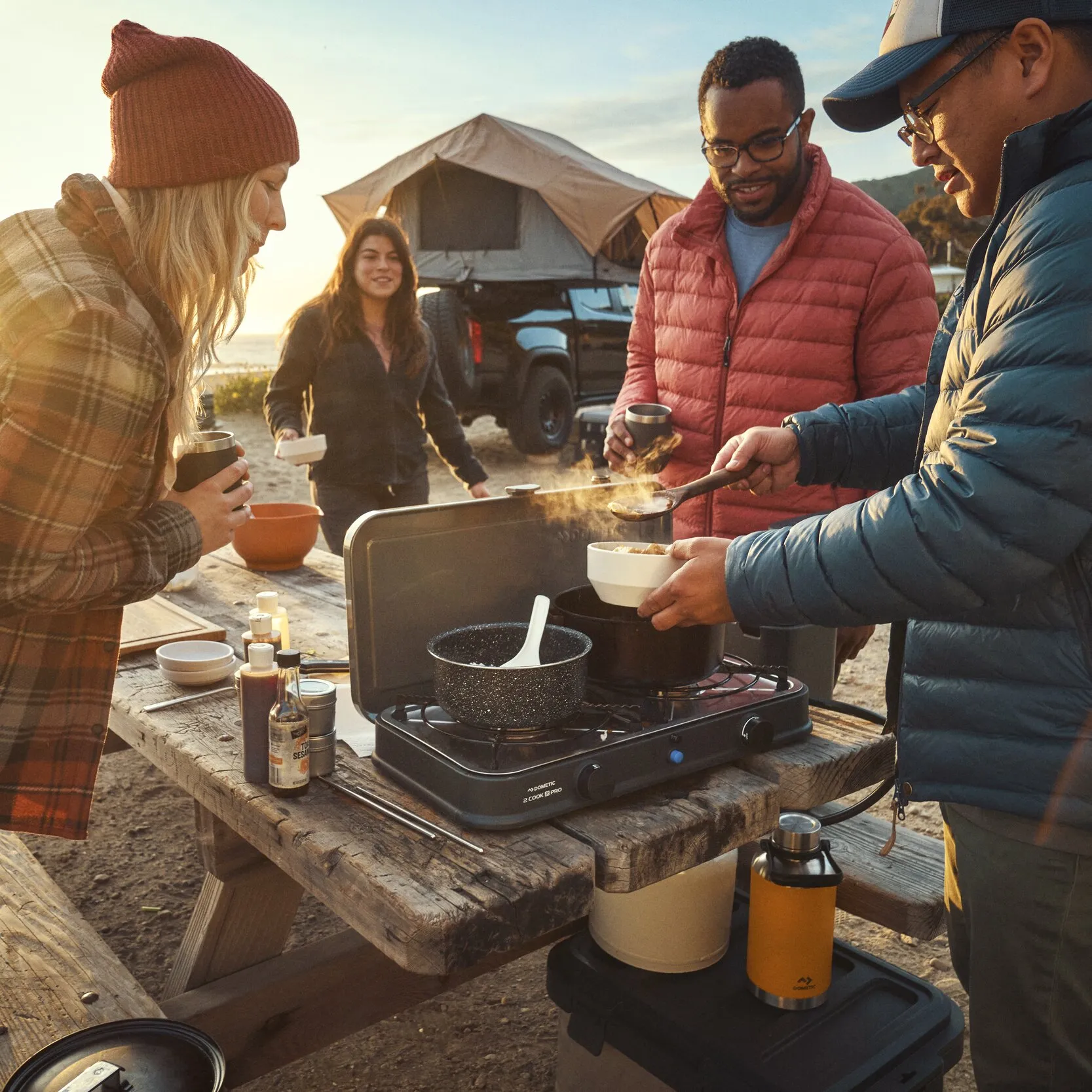 Vier Personen kochen am Campingkocher Van Cook FFD BBQ Plate auf einer Holztafel, neben ihnen gelbe Gasflasche, dampfender Topf und Becher, Zelt im Hintergrund
