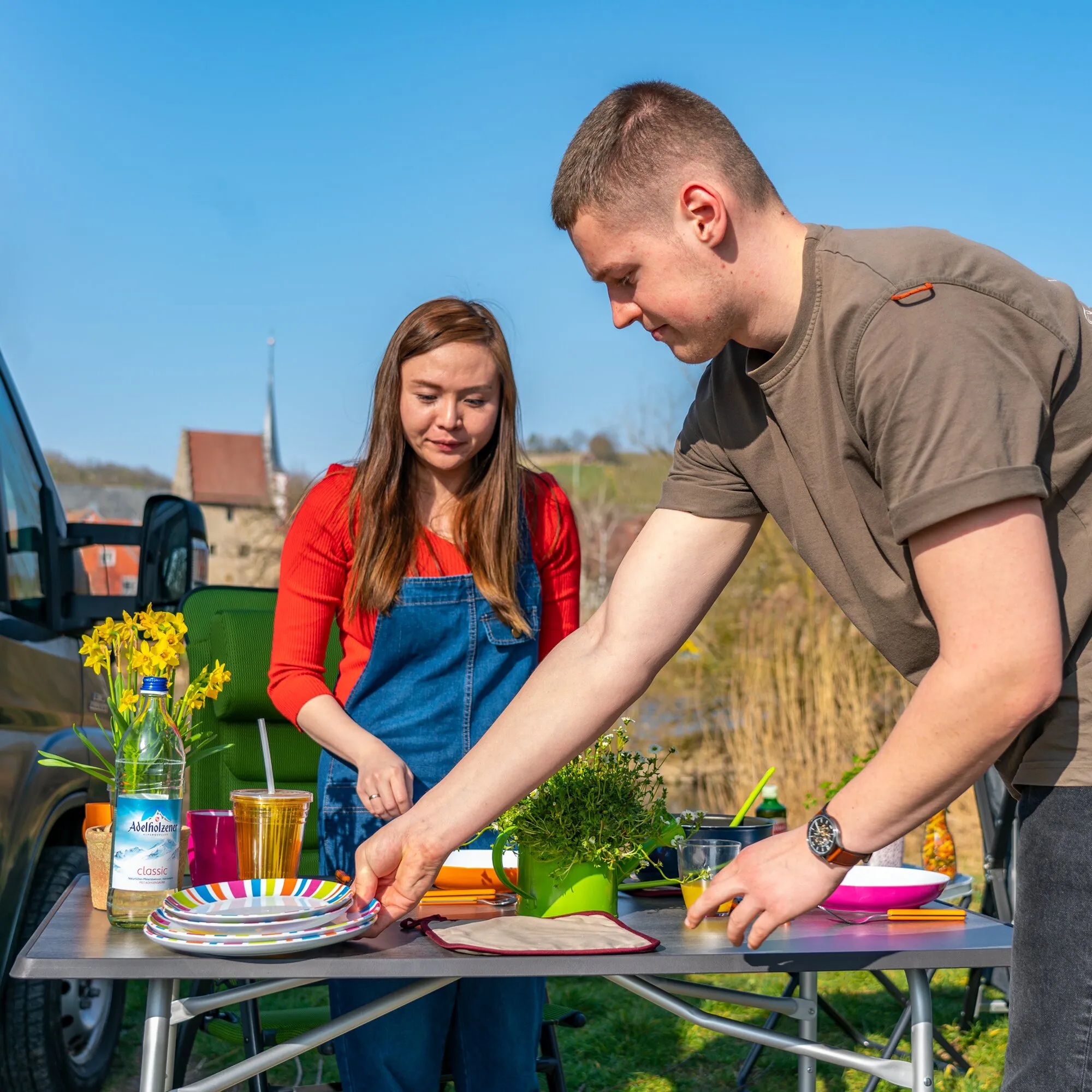 Grauer Campingtisch HighQ Blackline, 115 cm, im Freien mit Geschirr und Getränken; zwei Personen bereiten eine Mahlzeit vor