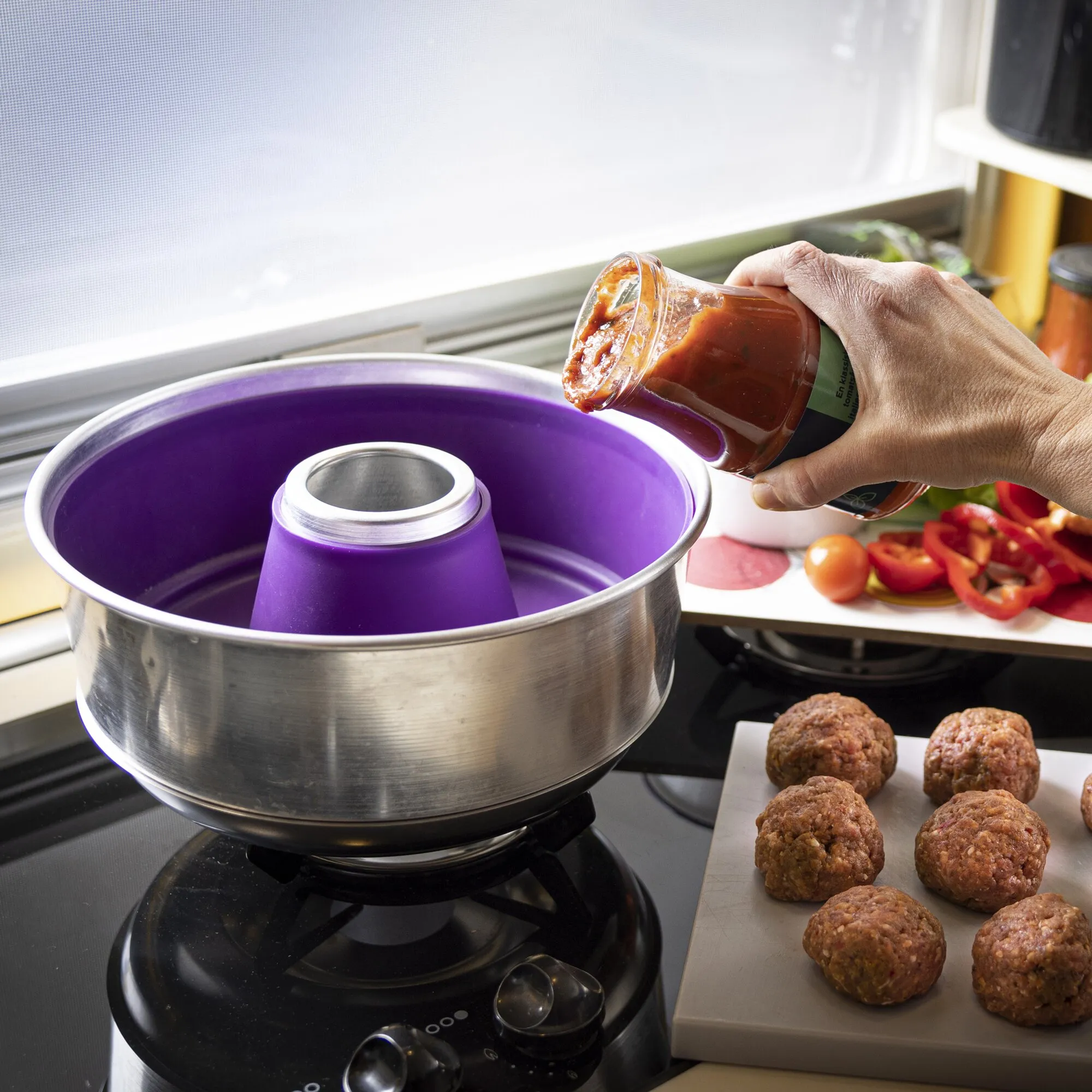 Violette Silikonform Maxi 3L mit Edelstahlrand auf dem Herd, während eine Hand Tomatensoße hineingießt; Fleischbällchen daneben