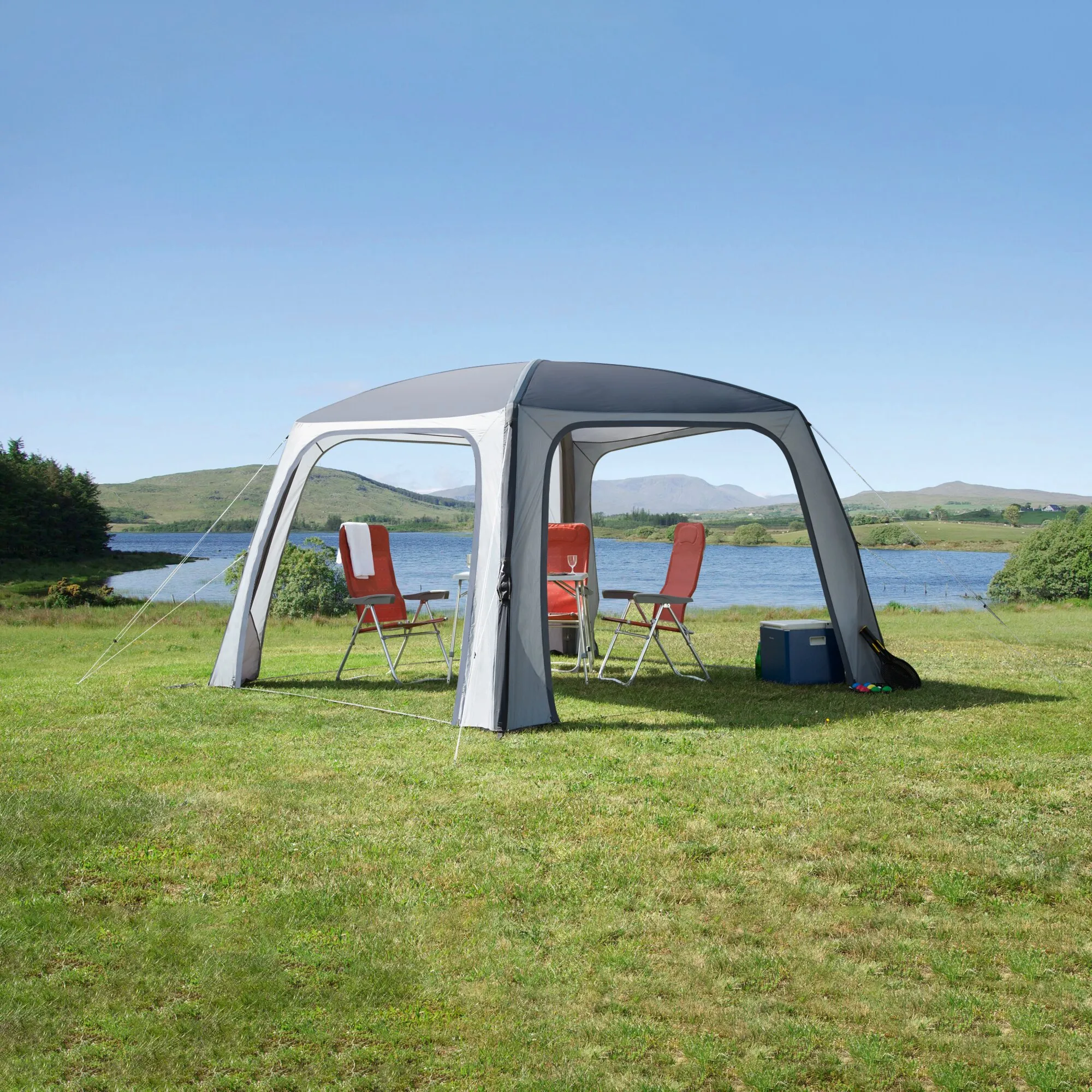 Grauer Pavillon Relax Air mit offenen Seiten, roten Campingstühlen und Tisch auf einer Wiese am Seeufer unerquicklich