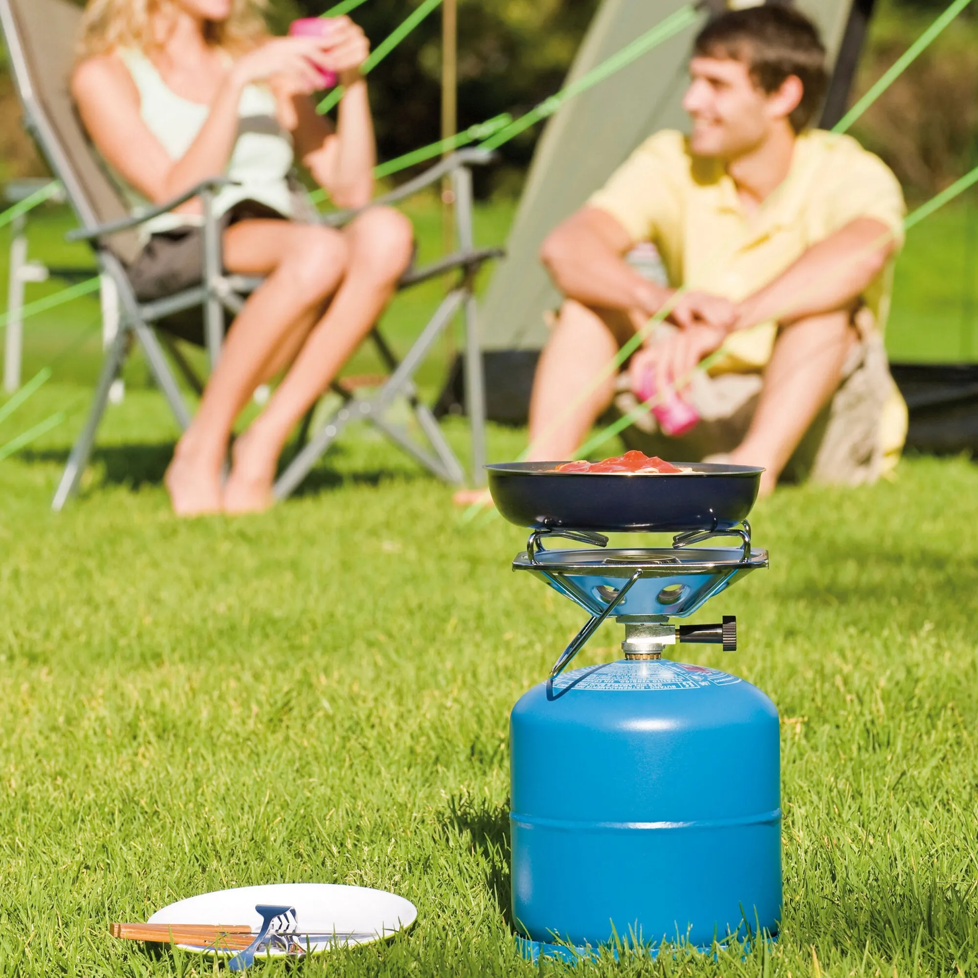 Campingkocher Super Carena R mit schwarzer Pfanne auf blauer Gasflasche beim Kochen auf einer Wiese vor einem Zeltpaar