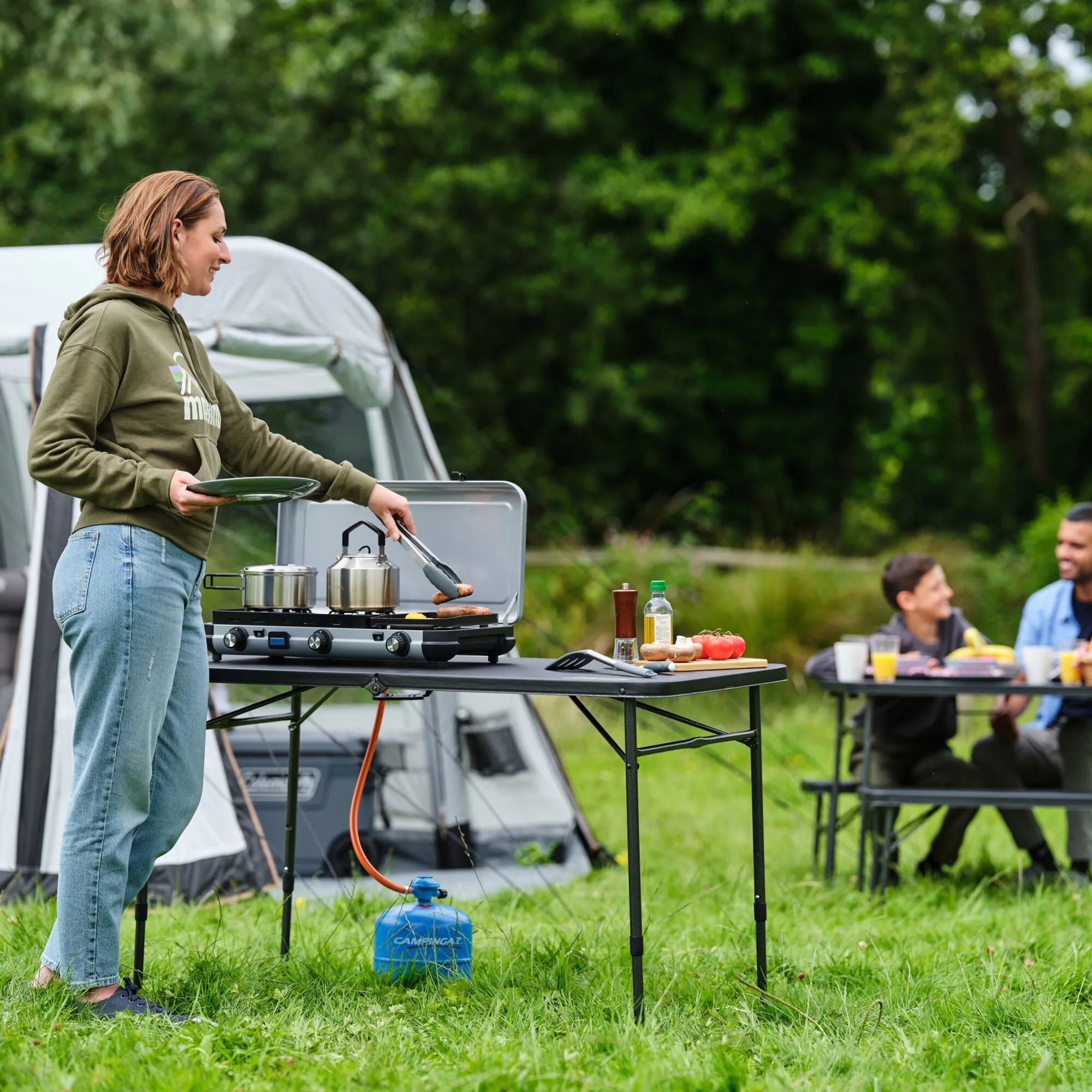 Camping-Gaskocher Camping Kitchen 2 MULTI-COOK mit zwei Töpfen und Grillplatte beim Kochen vor einem Zelt