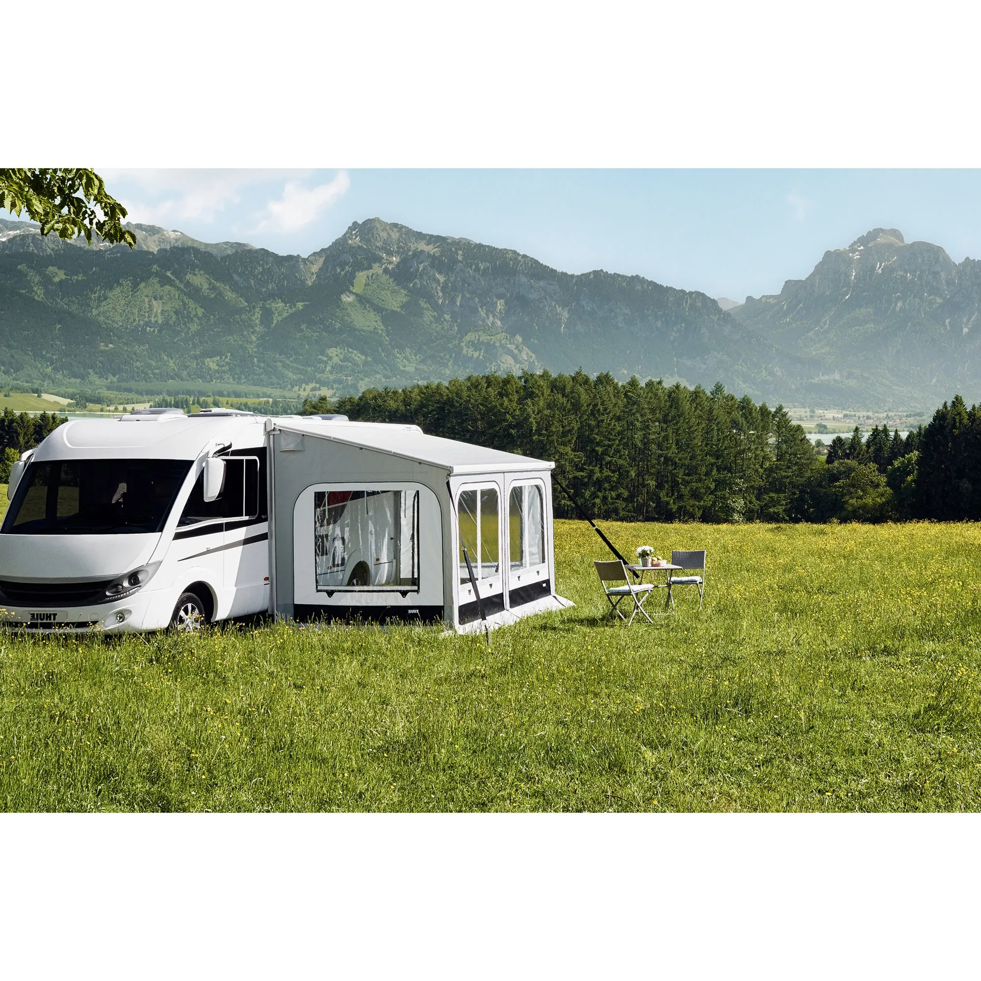 Weißes Wohnmobil mit aufgebautem grauem Markisenzelt Thule Panorama, Campingtisch und Stühlen auf einer Wiese vor Berglandschaft