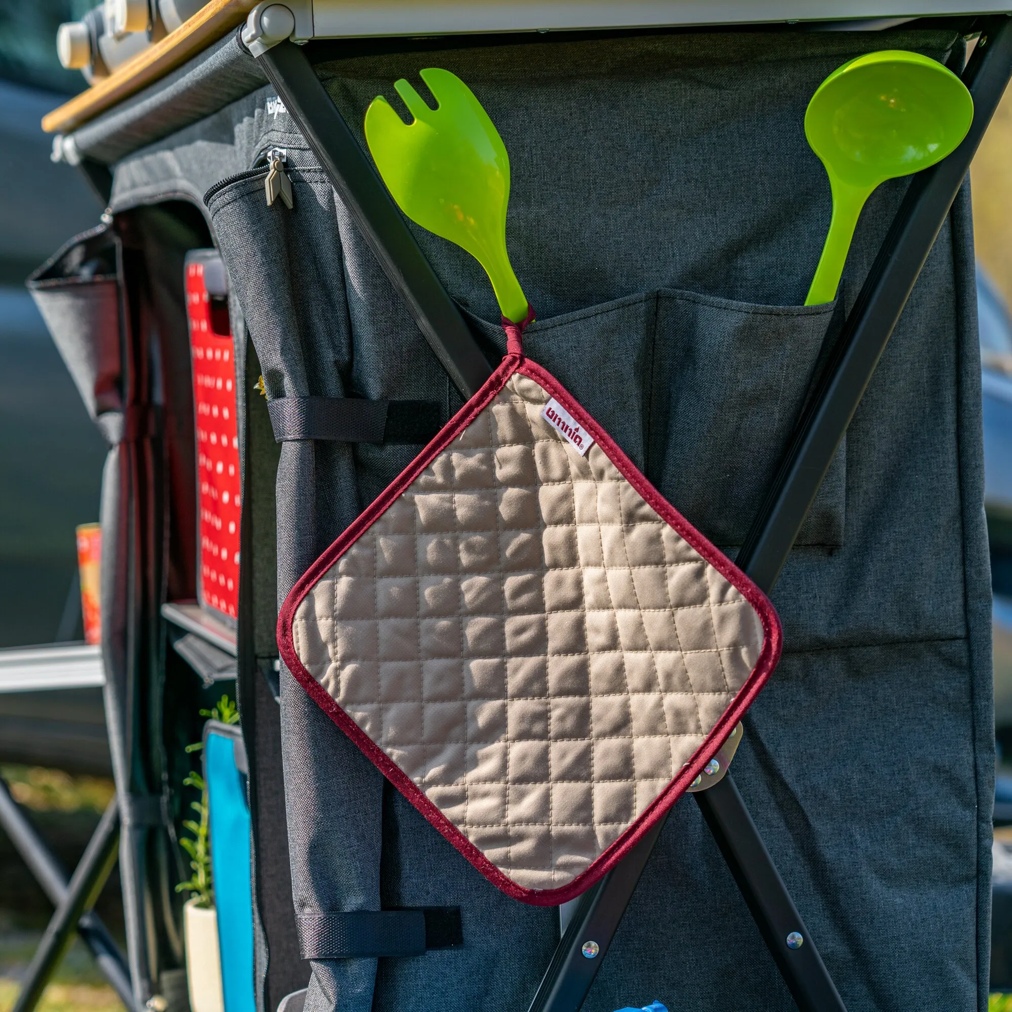 Beige karierter Topflappen mit roter Einfassung hängt an einer dunklen Küchen- oder Campingtasche neben grünen Küchenutensilien