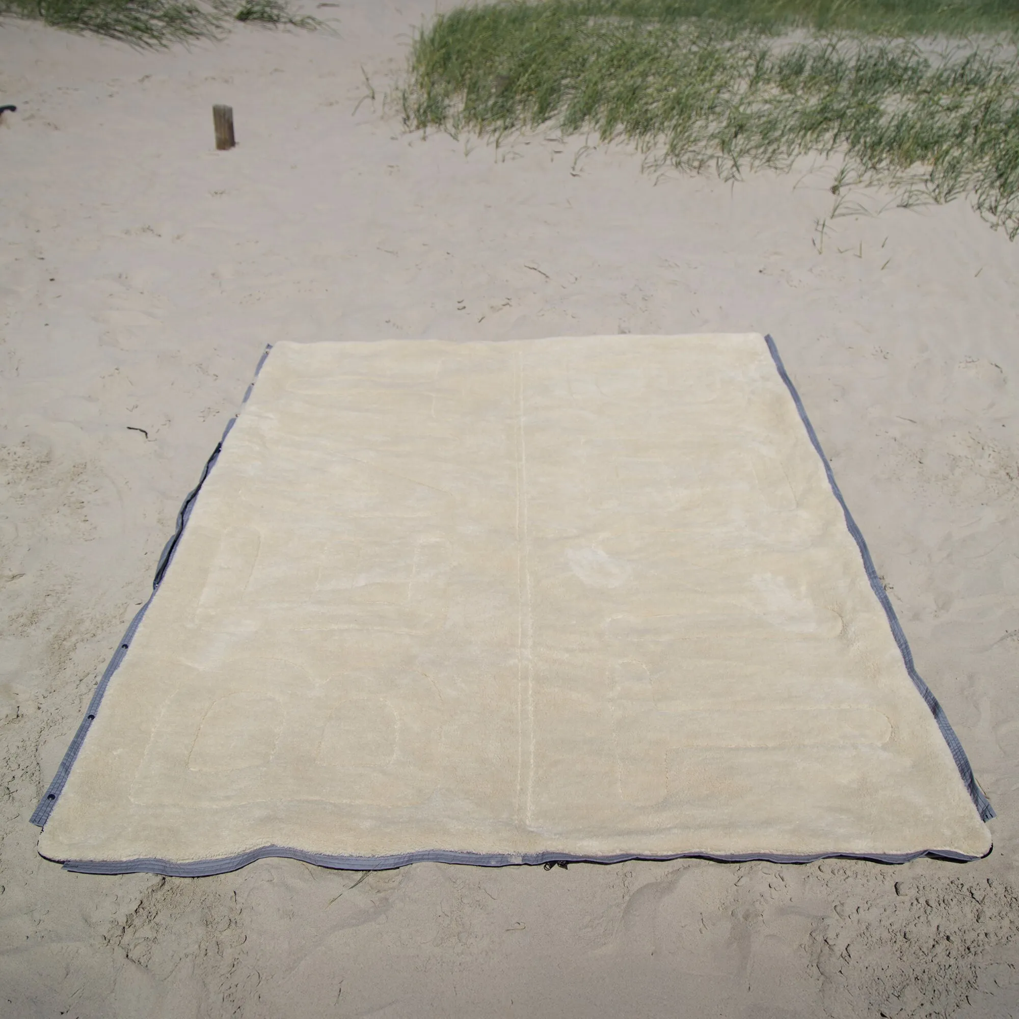 Auf Sand ausgebreiteter cremefarbener Vanlife-Schlafsack Bentcho mit grauem Rand und Reißverschluss, im Hintergrund Dünengras