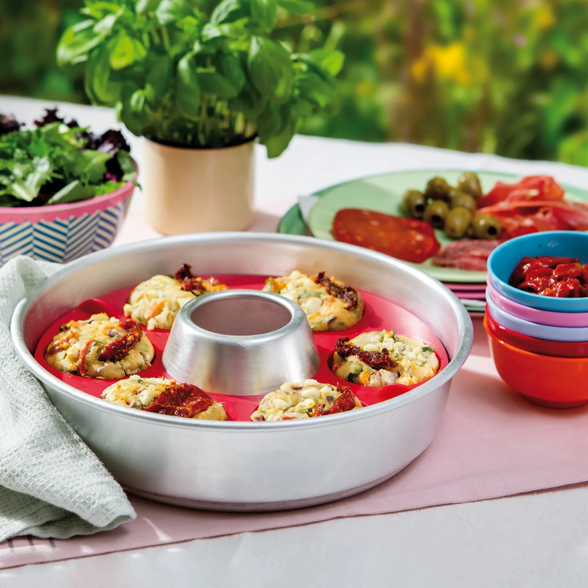 Muffin-Tab aus Metall mit roter Silikonform und herzhaften Muffins mit Käse und getrockneten Tomaten auf gedecktem Tisch