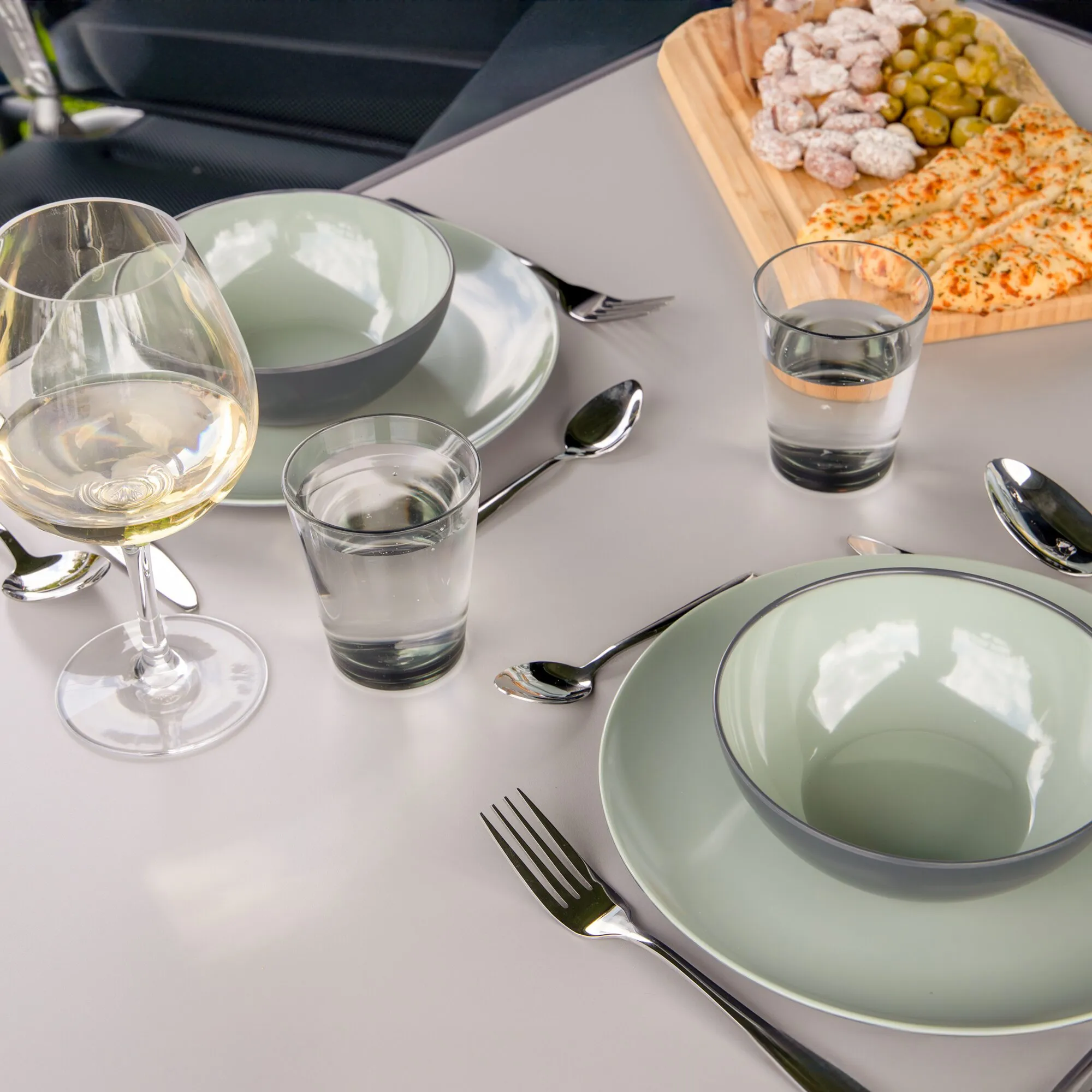 Hellgrünes Geschirrset Vivid Line mit Tellern und Schalen, gedeckter Tisch mit Gläsern, Besteck und Snacks auf einem Holzbrett